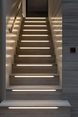 Modern staircase with built-in LED lights along each step, flanked by white walls and a gray wooden wall on the right.