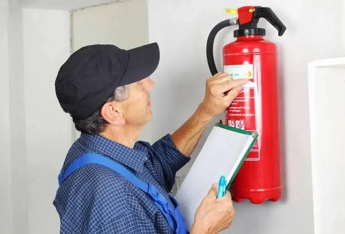 An older man in a black cap and blue checkered shirt inspecting a red fire extinguisher on a wall with a clipboard and pen.