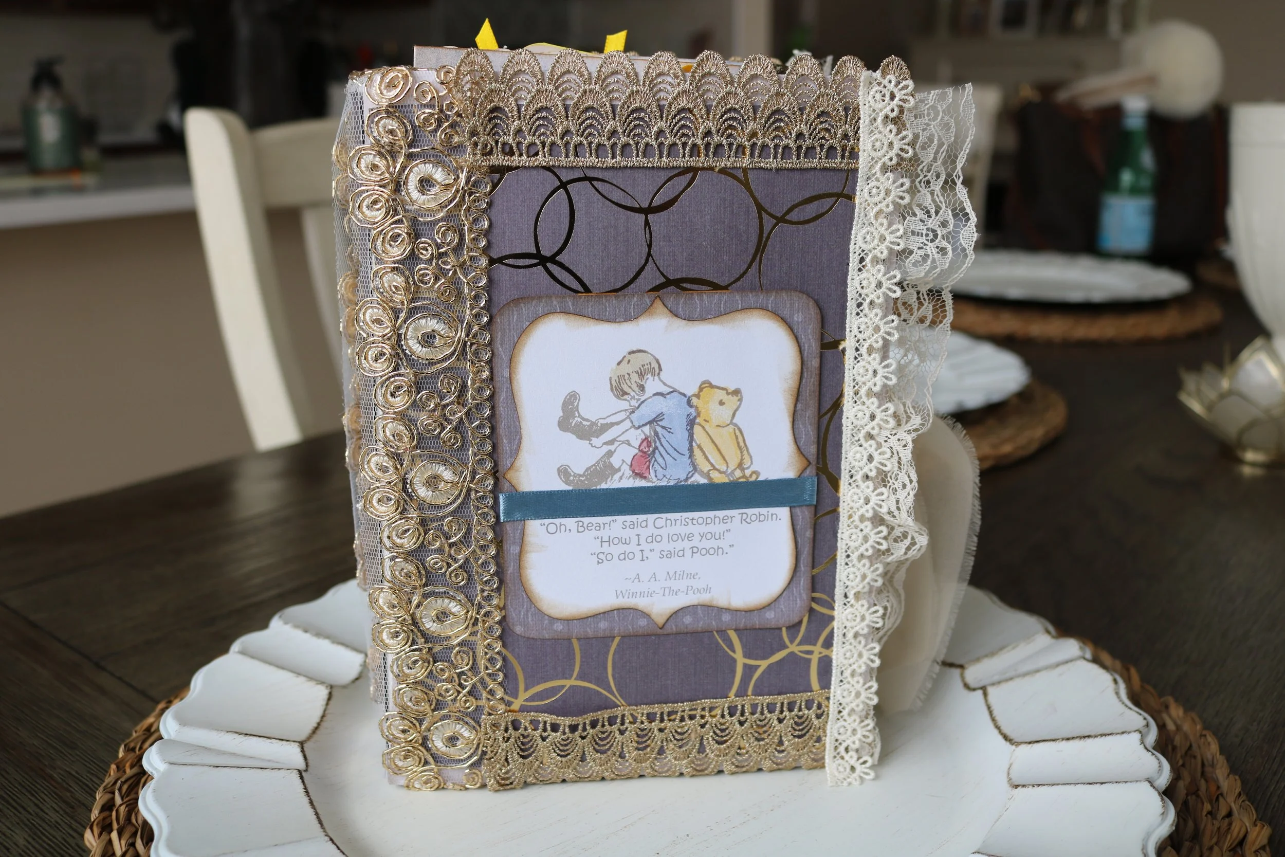 Decorative handmade journal with lace and gold embellishments, featuring a Winnie-the-Pooh quote with a small illustration of Pooh and Christopher Robin, placed on a white ceramic tray with a wicker border.