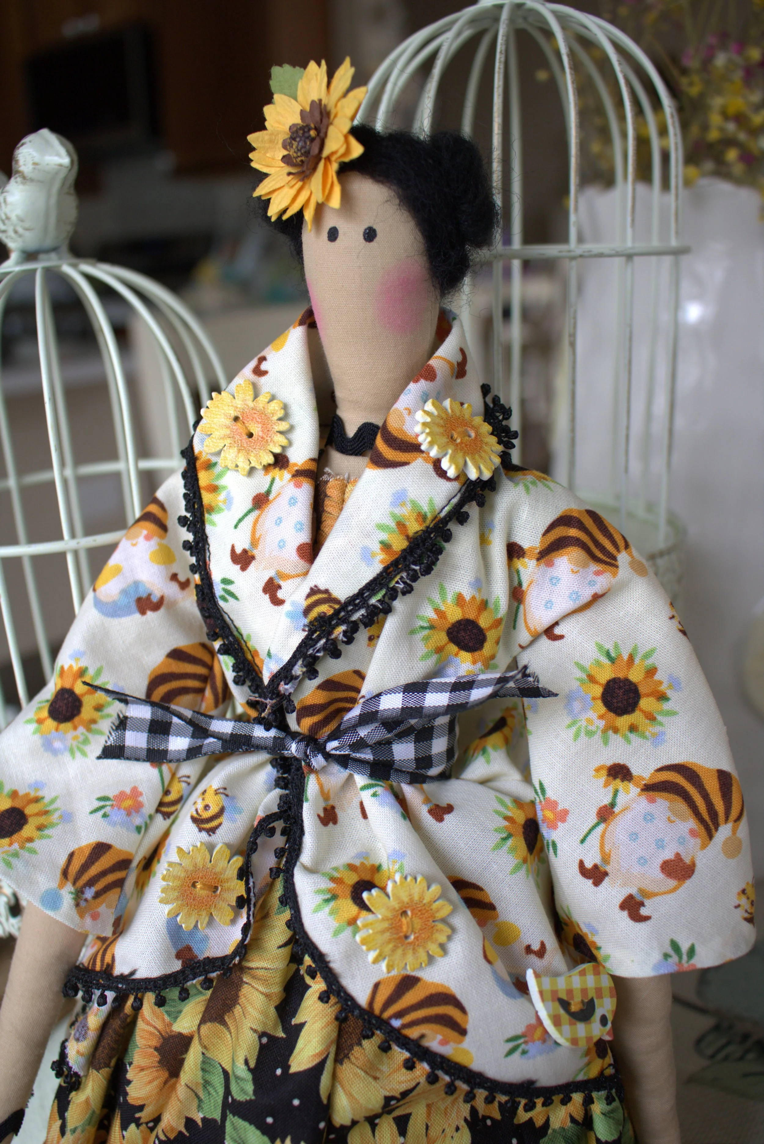 A handmade doll dressed in sunflower-themed clothing with a sunflower headpiece, black hair, and a beige face, positioned in front of two white birdcages.