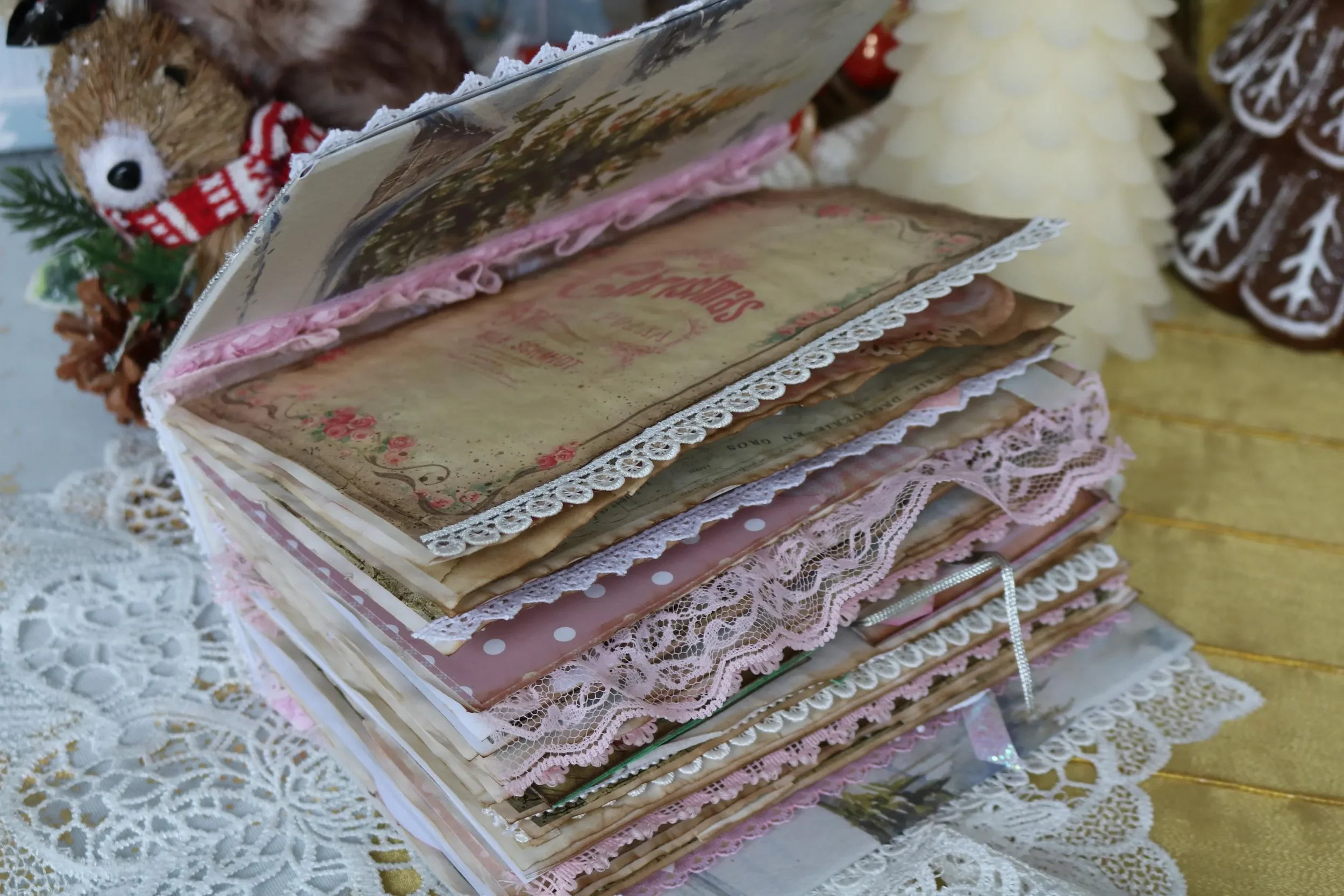 A decorated antique vintage junk journal with lace and frilled edges, on a lace tablecloth with Christmas-themed decorations in background.