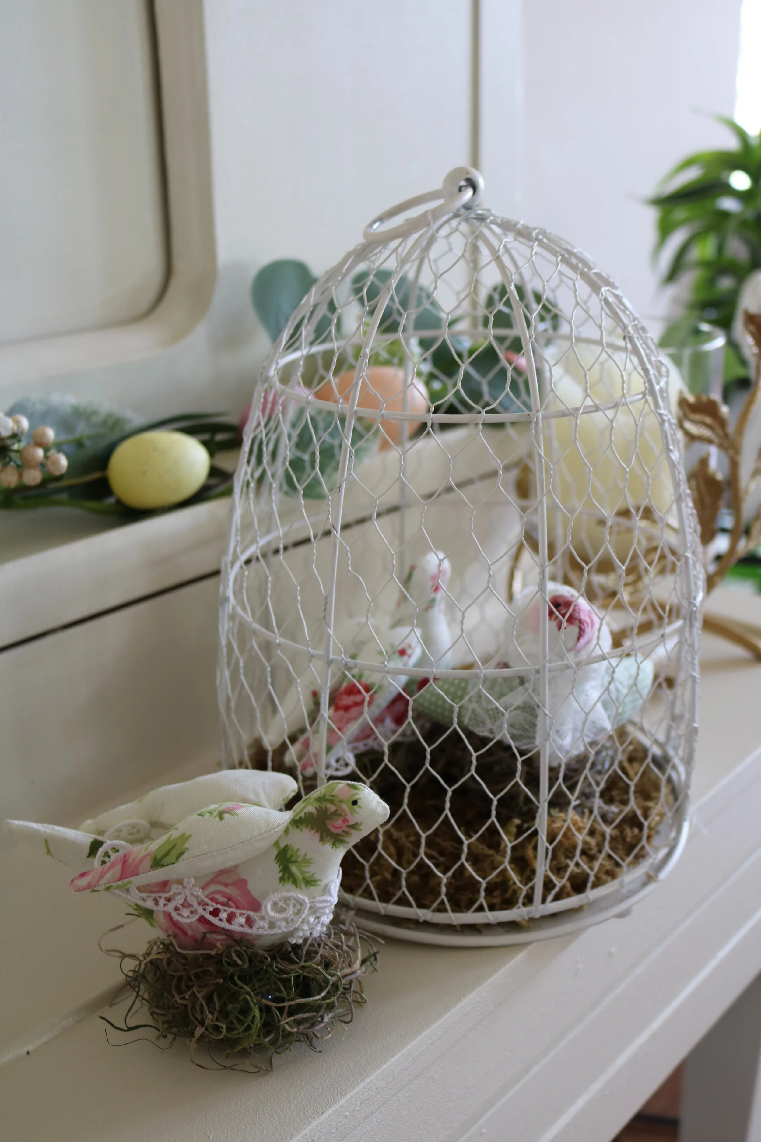 A decorative white wire birdcage with a small fabric sewn bird inside, placed on a white surface with surrounding Easter-themed decorations including a faux egg and a floral fabric. In the background, there are green plants and a mirror.