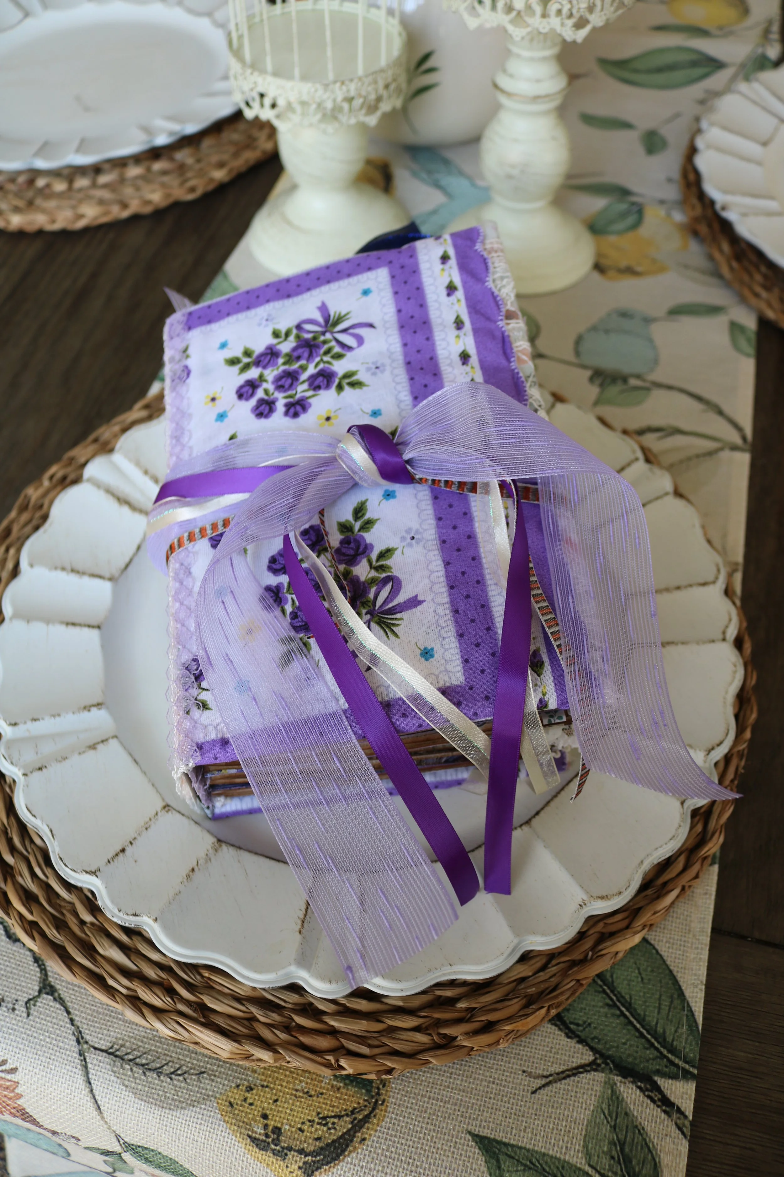 A lavender junk journal with a floral and purple design, purple ribbons, and a sheer purple bow, placed on a white decorative plate with a woven basket border, on a table with a floral tablecloth.