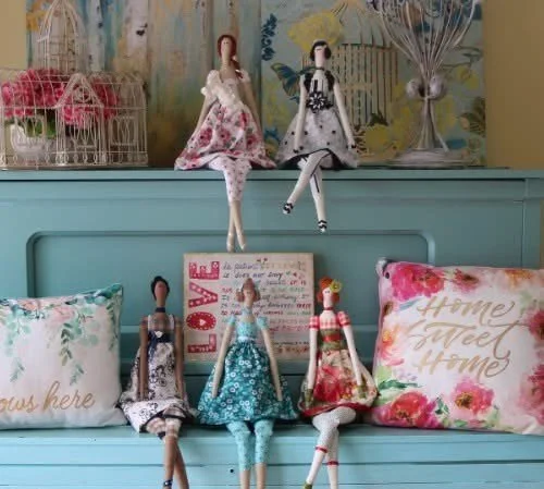 A decorative setup with five fabric dolls sitting on a blue surface, three dolls sitting on the surface and two dolls hanging on the wall. There are floral pillows, one with the text "Home Sweet Home" and another with floral patterns. The background features a painted wall with bird and flower designs, and a wire basket with pink flowers.