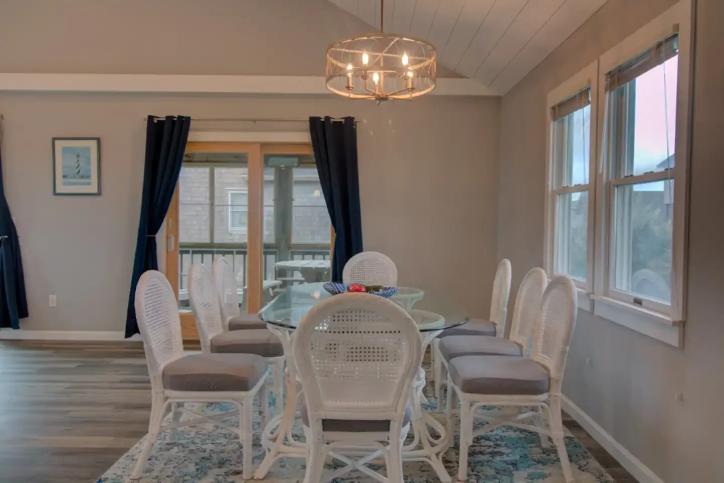 Dining room with a glass-top table surrounded by  white wicker chairs with beige cushions. There is a chandelier hanging from the ceiling, and a sliding glass door with navy curtains leading to a balcony. Three windows with beige blinds are on the ri