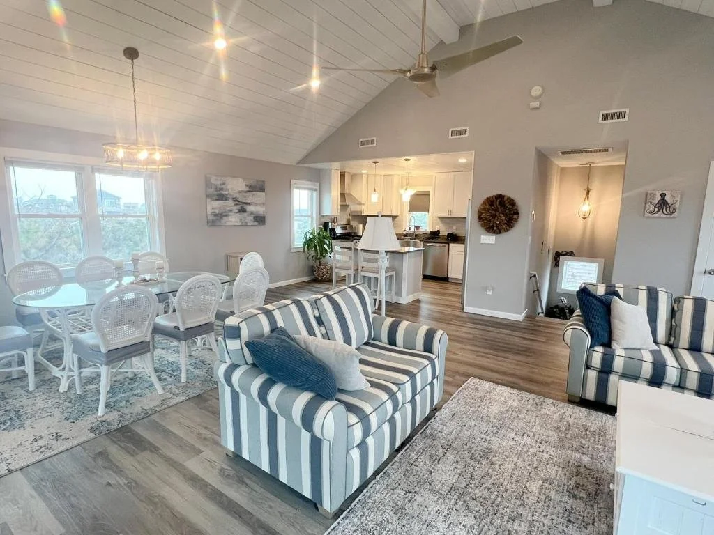 Open-concept living room and dining area with a view of the kitchen. Features striped sofas, a dining table with white chairs, and decorative wall art. Large windows let in natural light.