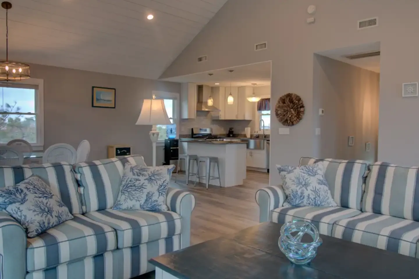 Living room with striped beige and blue sofas, decorative pillows, a coffee table with glass art, next to an open kitchen with white cabinets, bar stools, and pendant lighting.