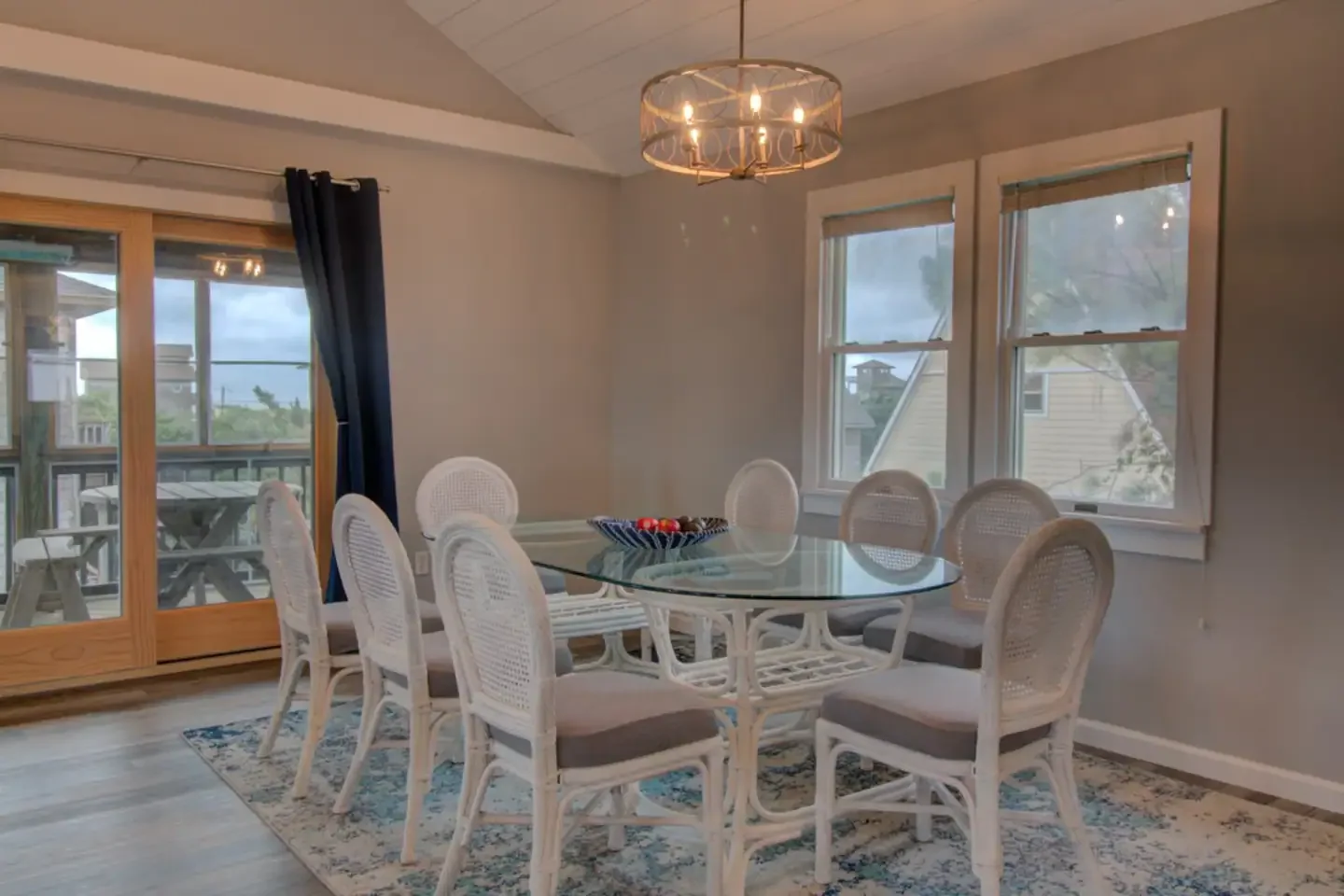 Dining room with a glass-top table surrounded by eight white cane-back chairs with beige cushions, a patterned rug underneath, a chandelier overhead, large windows, and a sliding door leading to a screened porch with outdoor seating.