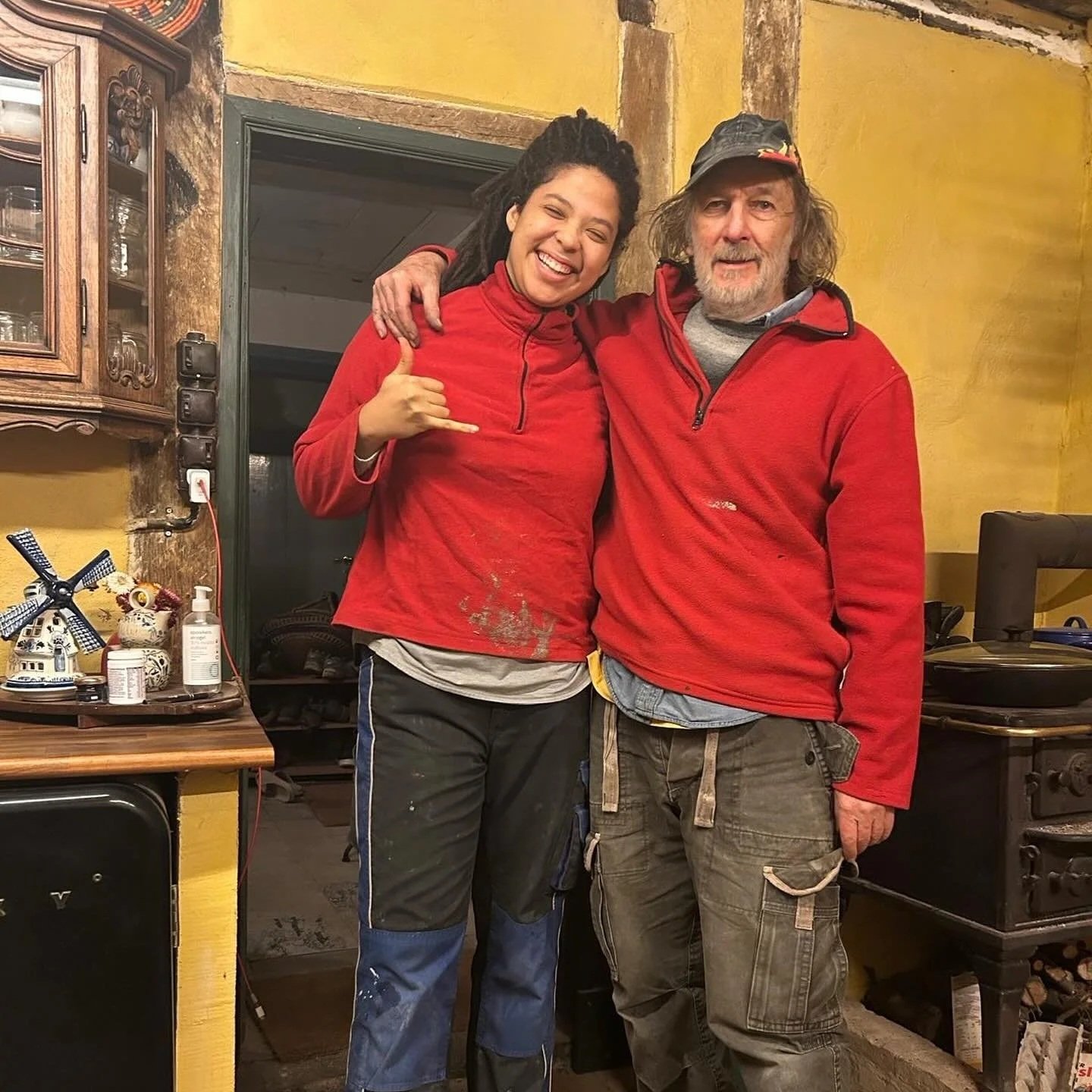 Two people hugging inside a cozy, rustic kitchen. The woman on the left is smiling, with dreadlocks, wearing a red jacket and black pants. The man on the right has a beard and is wearing a red fleece jacket, gray shirt, and a baseball cap. The background shows kitchen shelves, a stove, and various decorative items.
