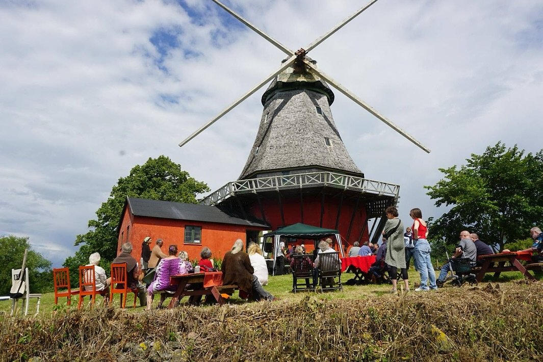 A group of people gathered outdoors near a historic windmill with large blades, a conical roof, and a red lower section, under cloudy skies.