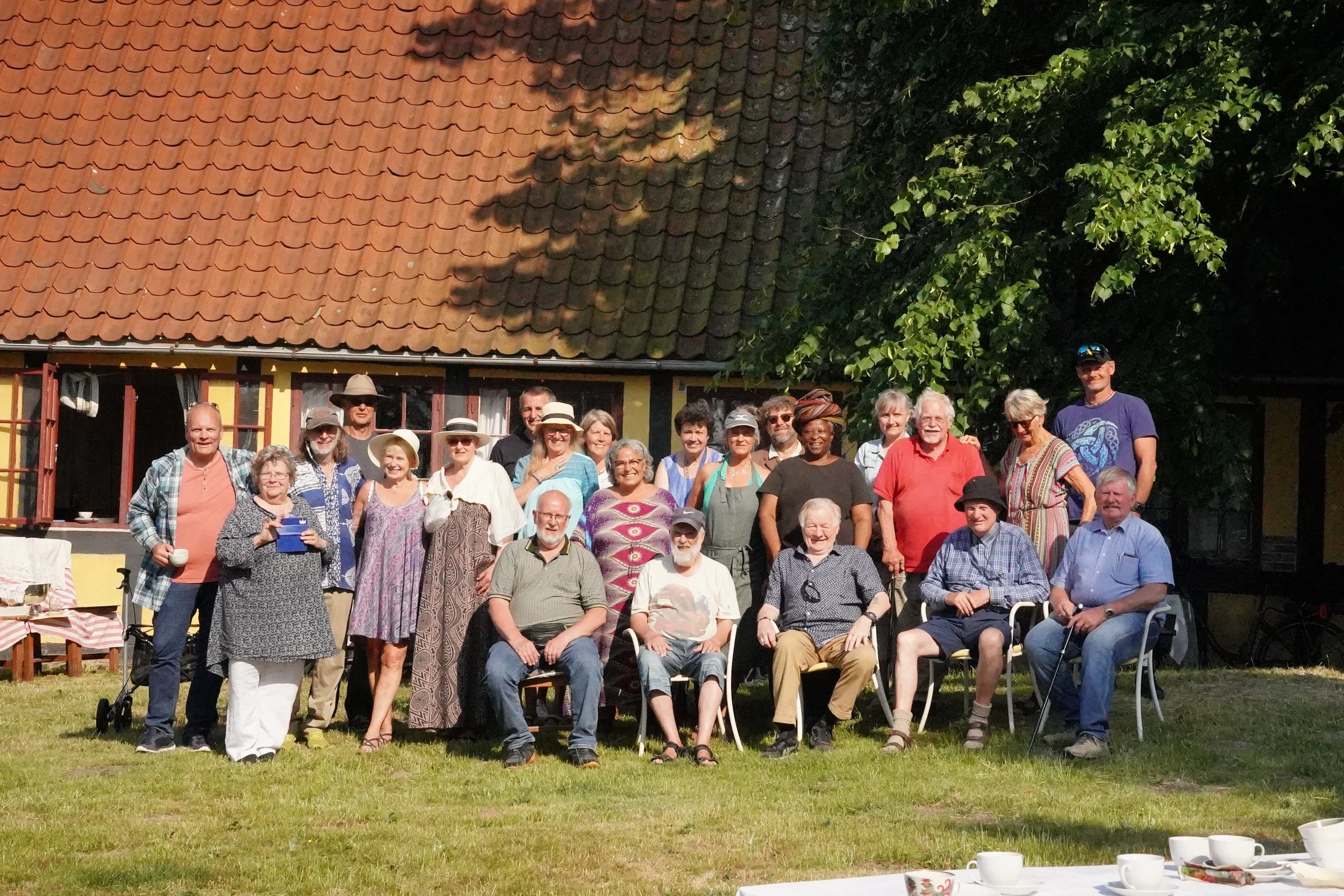 A large group of elderly people gathered outdoors in front of a house for a social event, with some sitting and some standing, and tea cups on a table in the foreground.