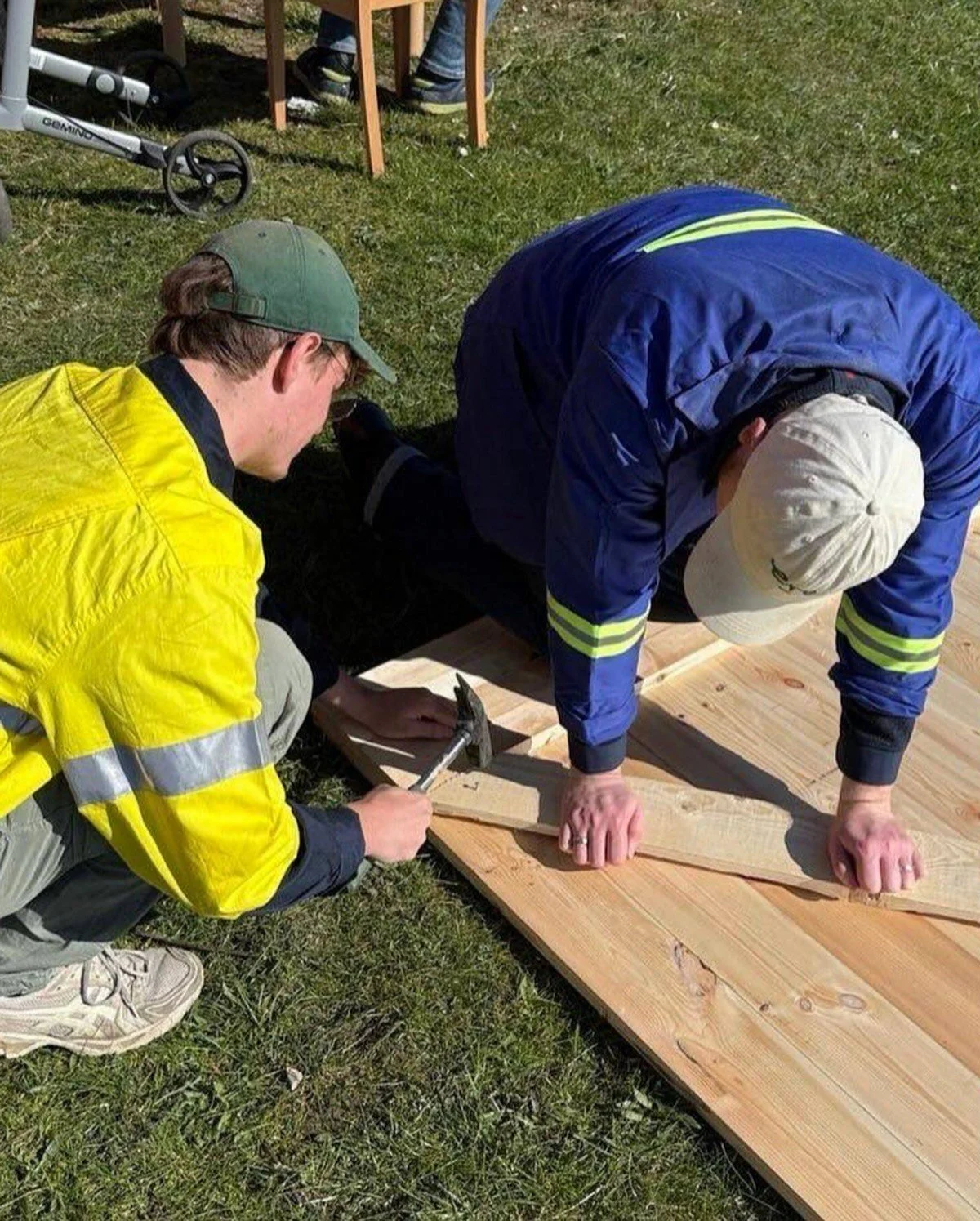 Two men working together on a wooden piece outdoors, with one holding a hammer and the other supporting the wood, on a grassy area.