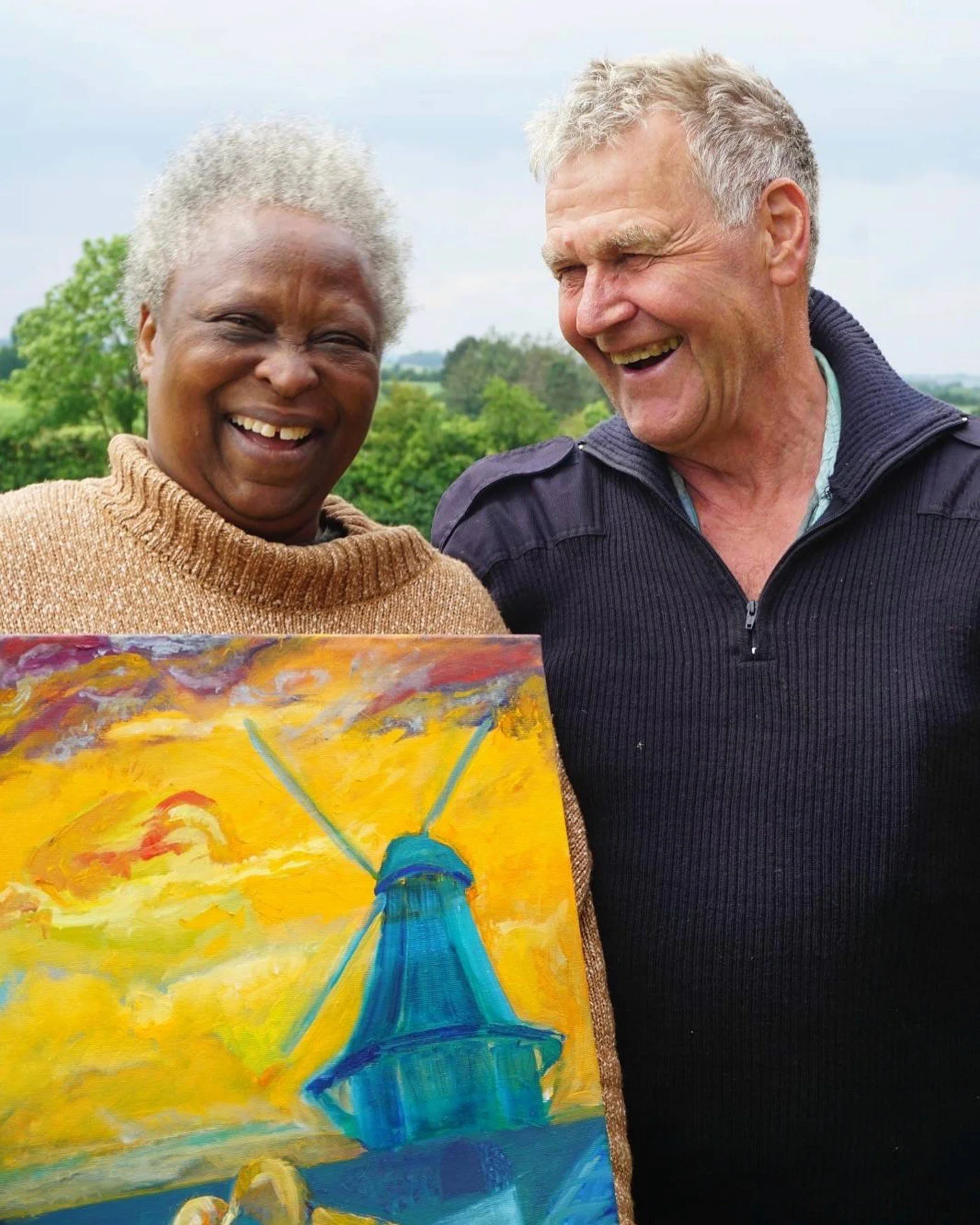 Two elderly people, a woman and a man, laughing outdoors during daytime, with a painting of a bridge and yellow sky in the foreground.