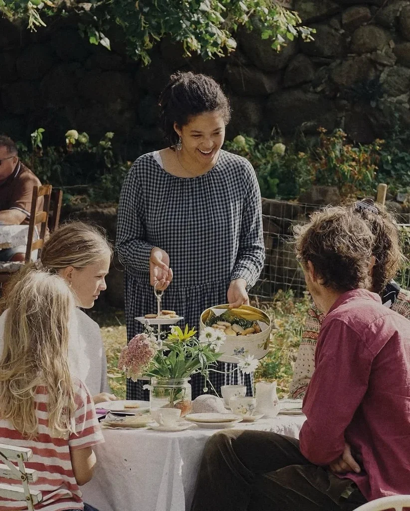 A woman in a checkered dress is serving food to people sitting at a table outdoors during daytime.