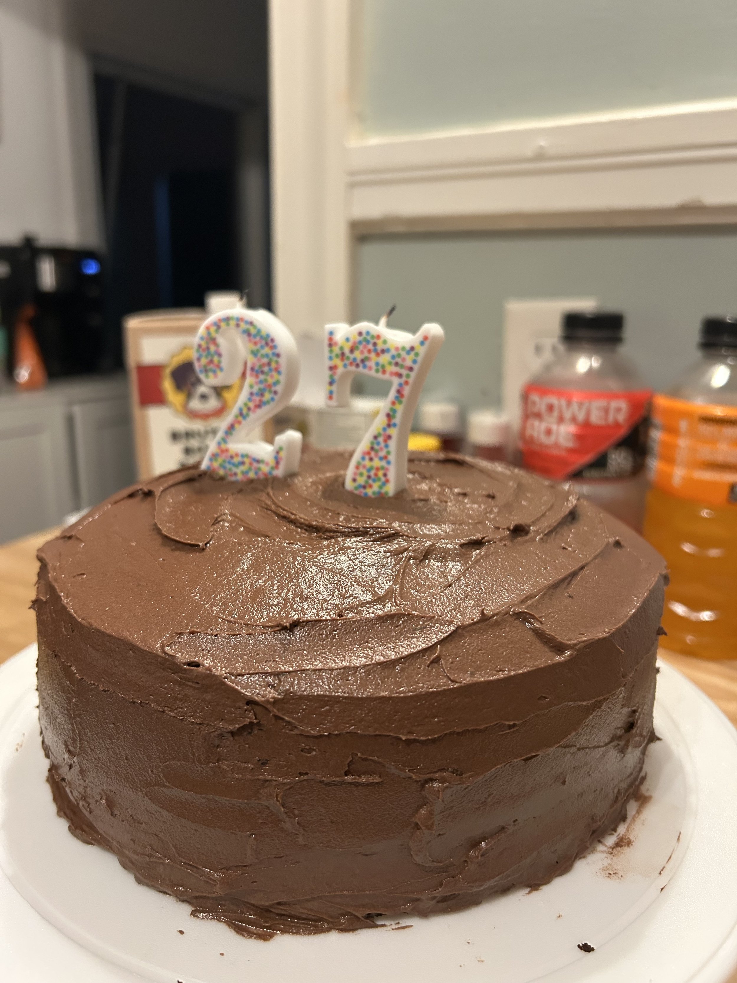 Chocolate cake with two lit birthday candles shaped as the numbers 2 and 7, decorated with multicolored sprinkles.