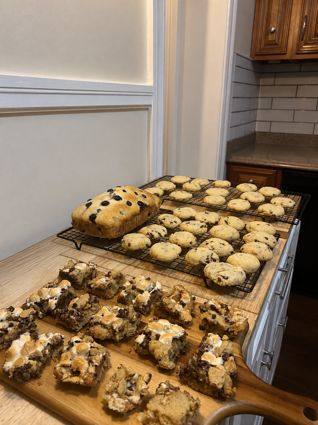 Freshly baked chocolate chip cookies and blondies cooling on racks and a cutting board on a kitchen counter.