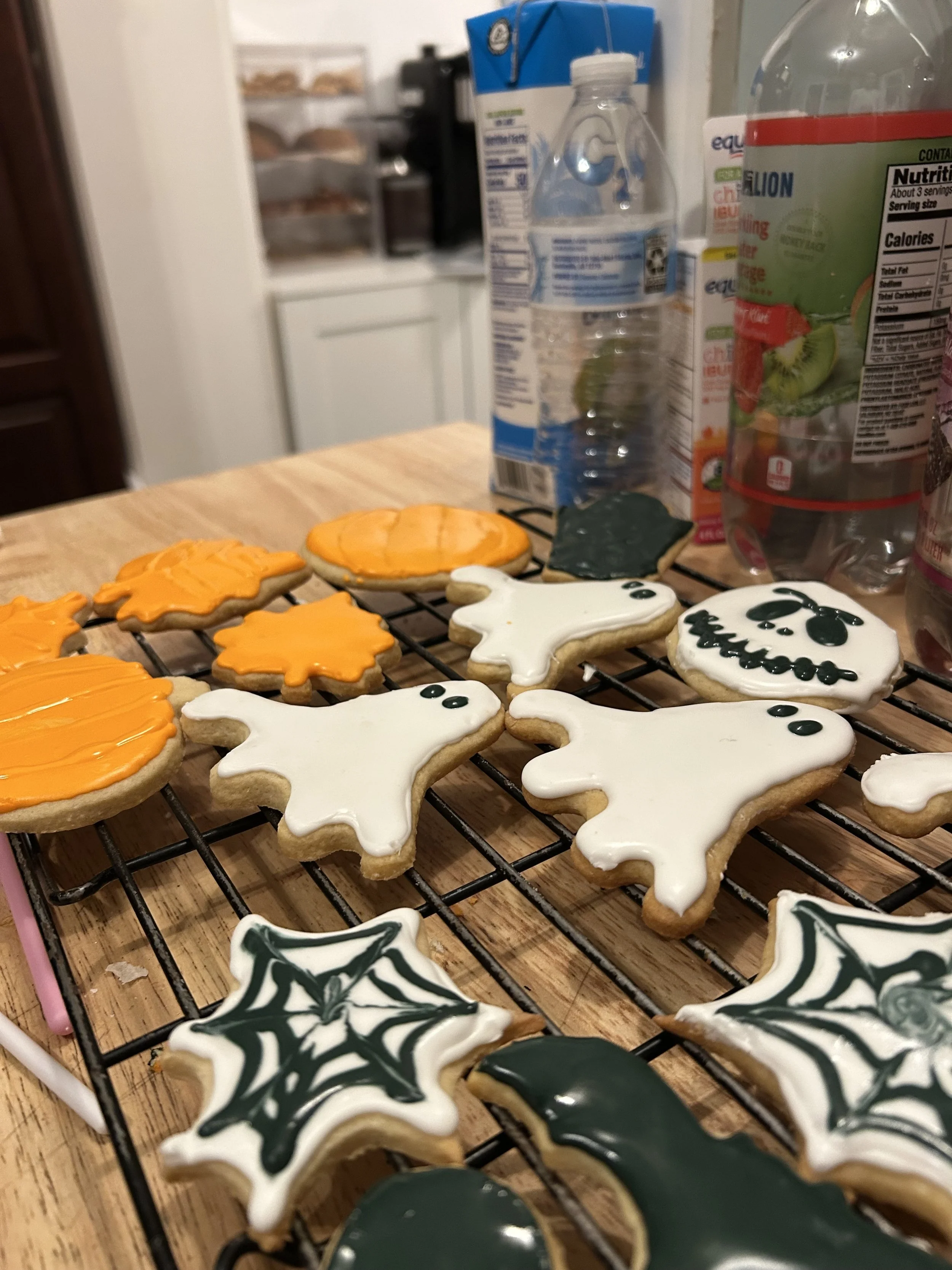 Halloween-themed cookies decorated with orange, white, and black icing, placed on a cooling rack on a wooden table, with a kitchen in the background.