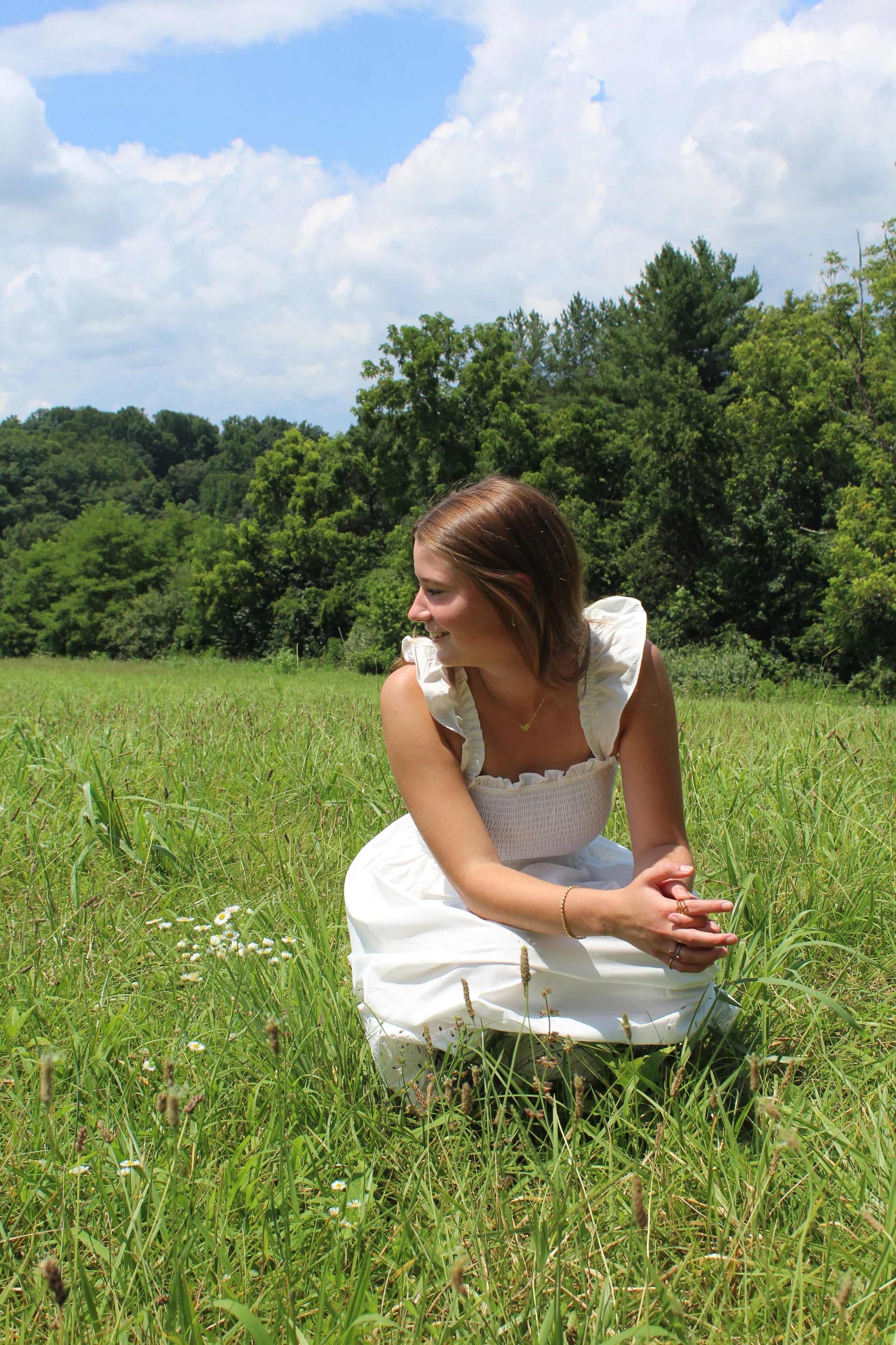 A woman in a white dress sitting in a grassy field during the day, with trees and clouds in the background.