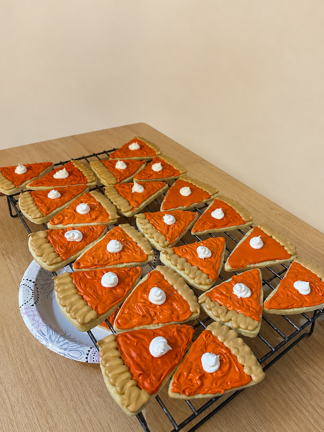Sugar cookies decorated as mini slices of pie with orange icing, beige border, and dollops of white icing on top, cooling on a wire rack.