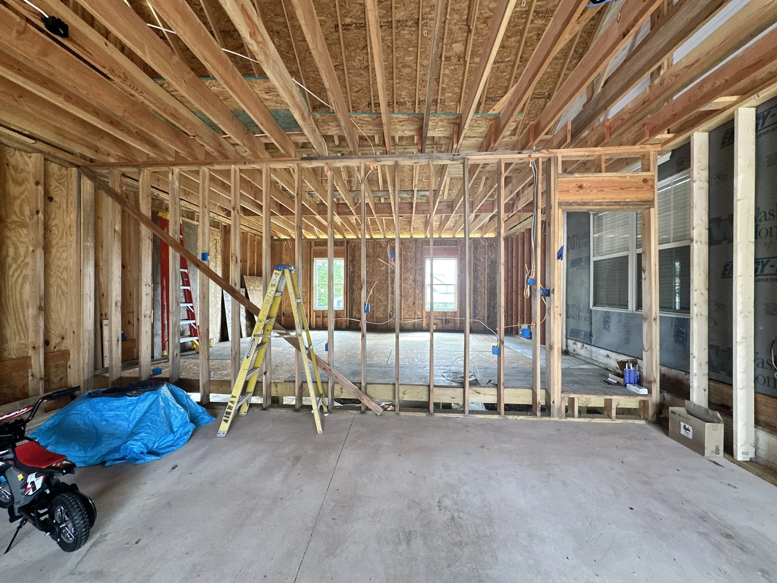 Interior of a house under construction with exposed wooden framing, electrical wiring, and windows. Construction tools and materials are visible, including ladders, a blue tarp, and a child's tricycle.