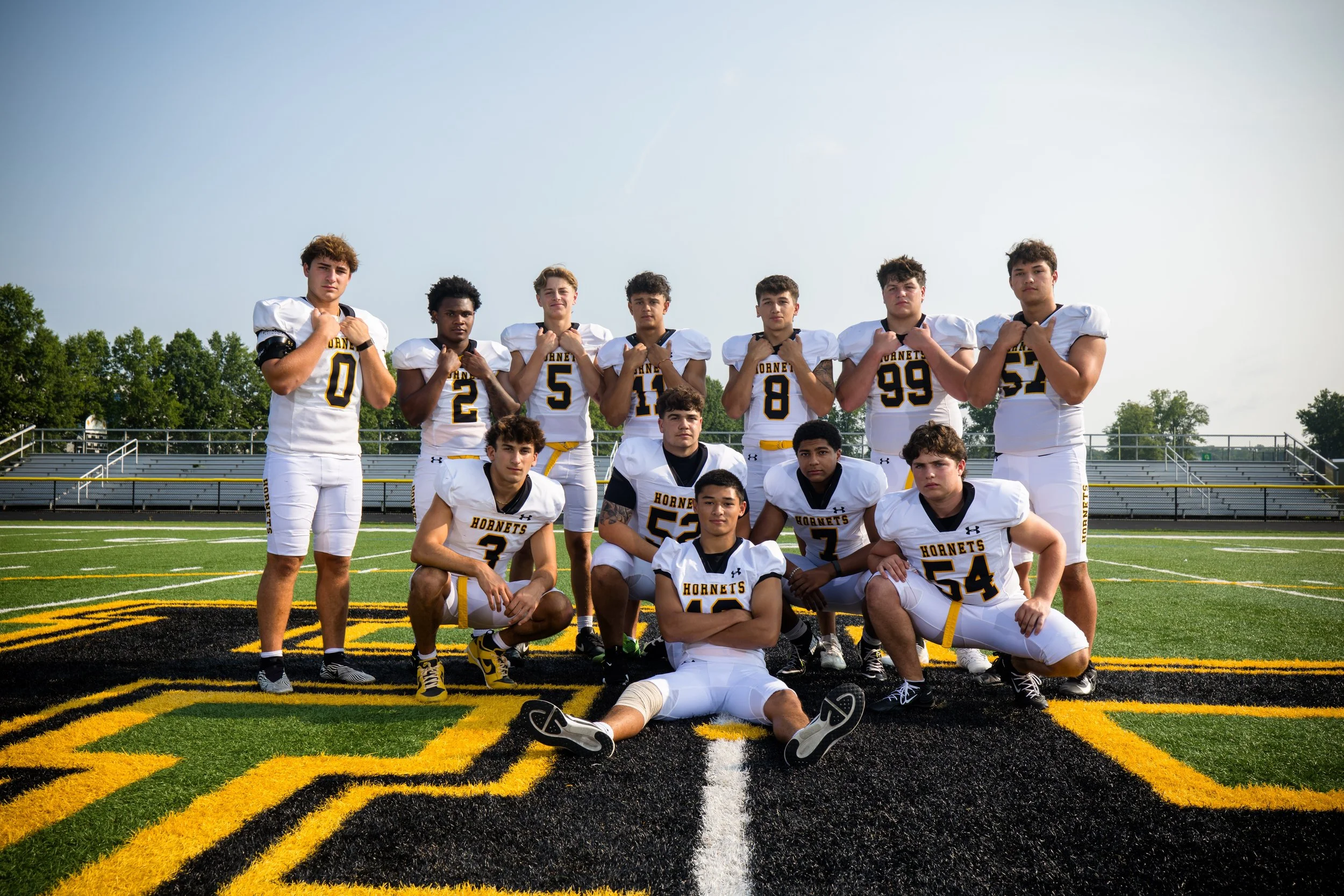 A group of high school football players in uniform posing on a football field with a track and trees in the background.