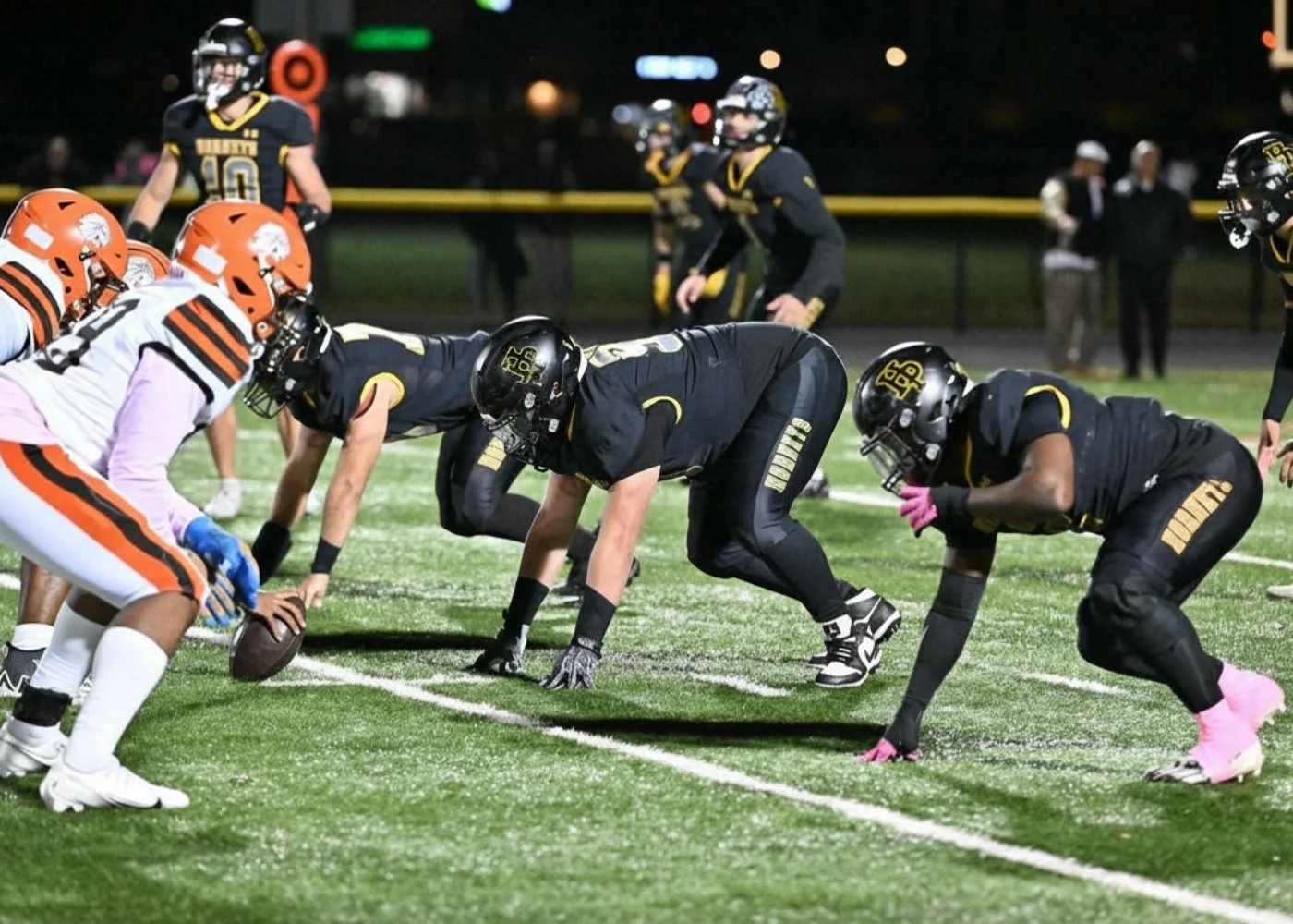 American football game with players in formation at the line of scrimmage, wearing black and white uniforms, on a lit field at night.