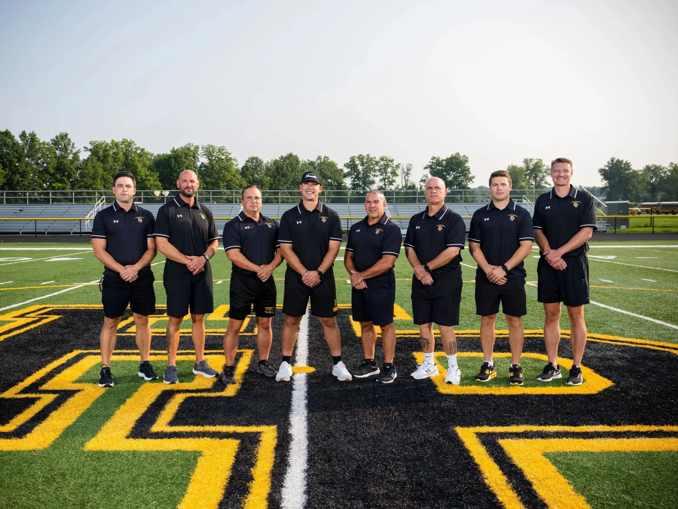 A group of eight men standing on a football field, wearing black and gold athletic shirts, with a school logo, and standing shoulder to shoulder in front of bleachers and trees.