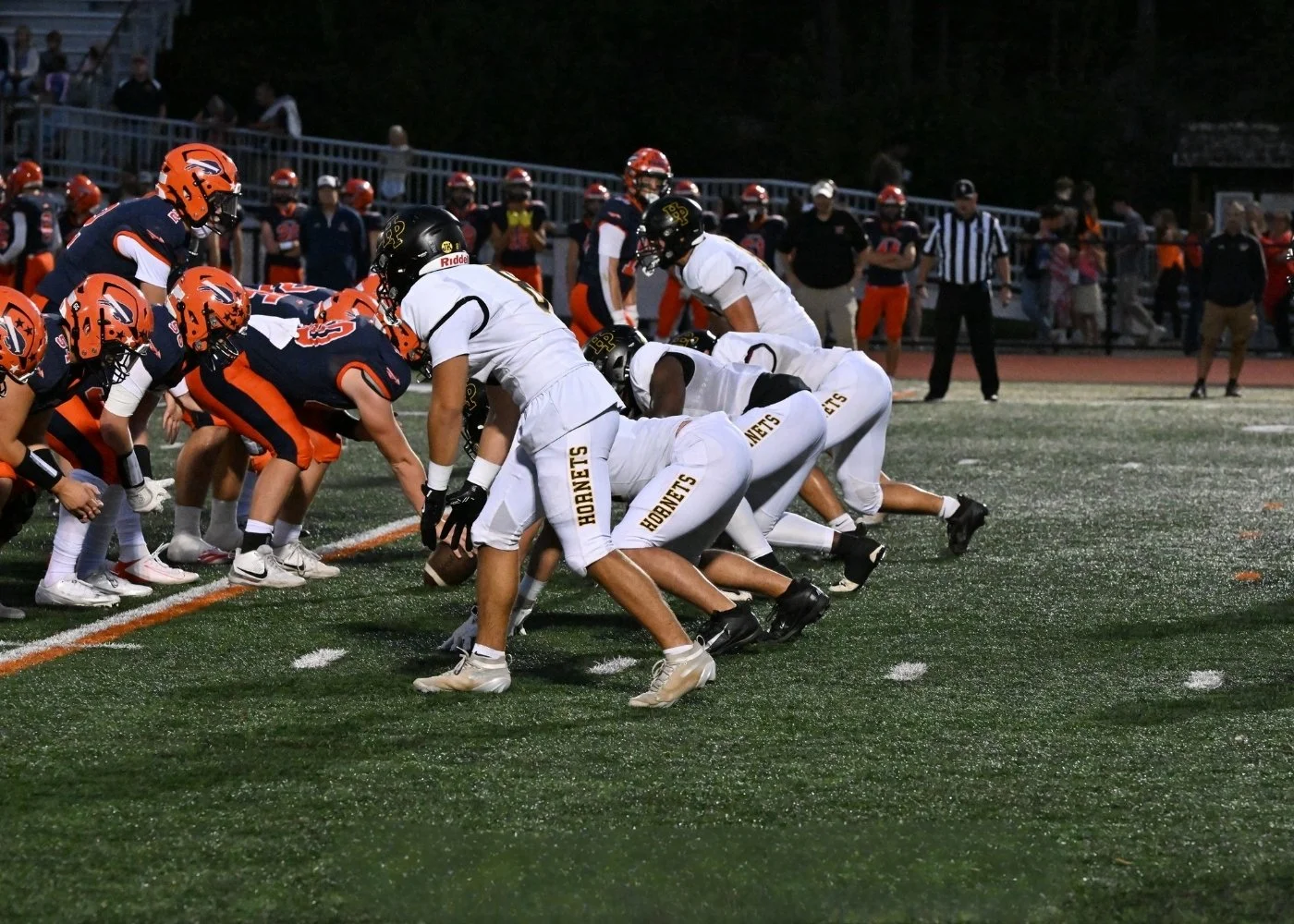 American football game with players in white and dark uniforms lined up at the line of scrimmage on a field during evening.