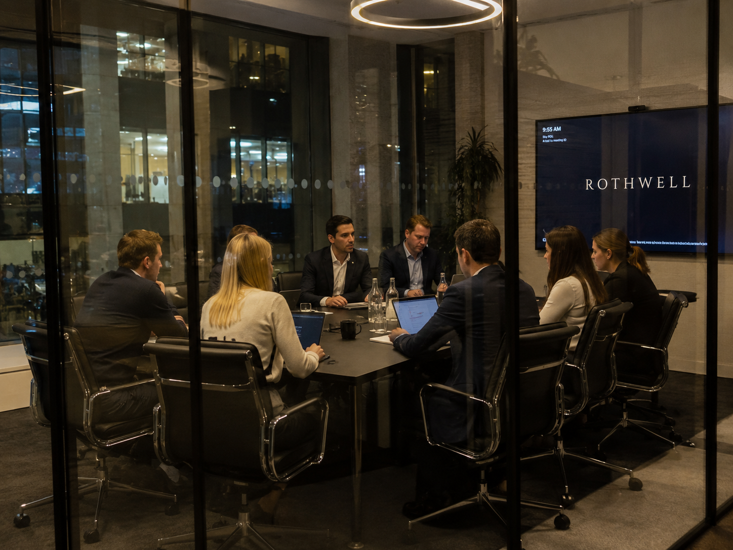 Business meeting through glass wall at conference room with nine people, laptops, water bottles, and a Rothwell display on the wall.