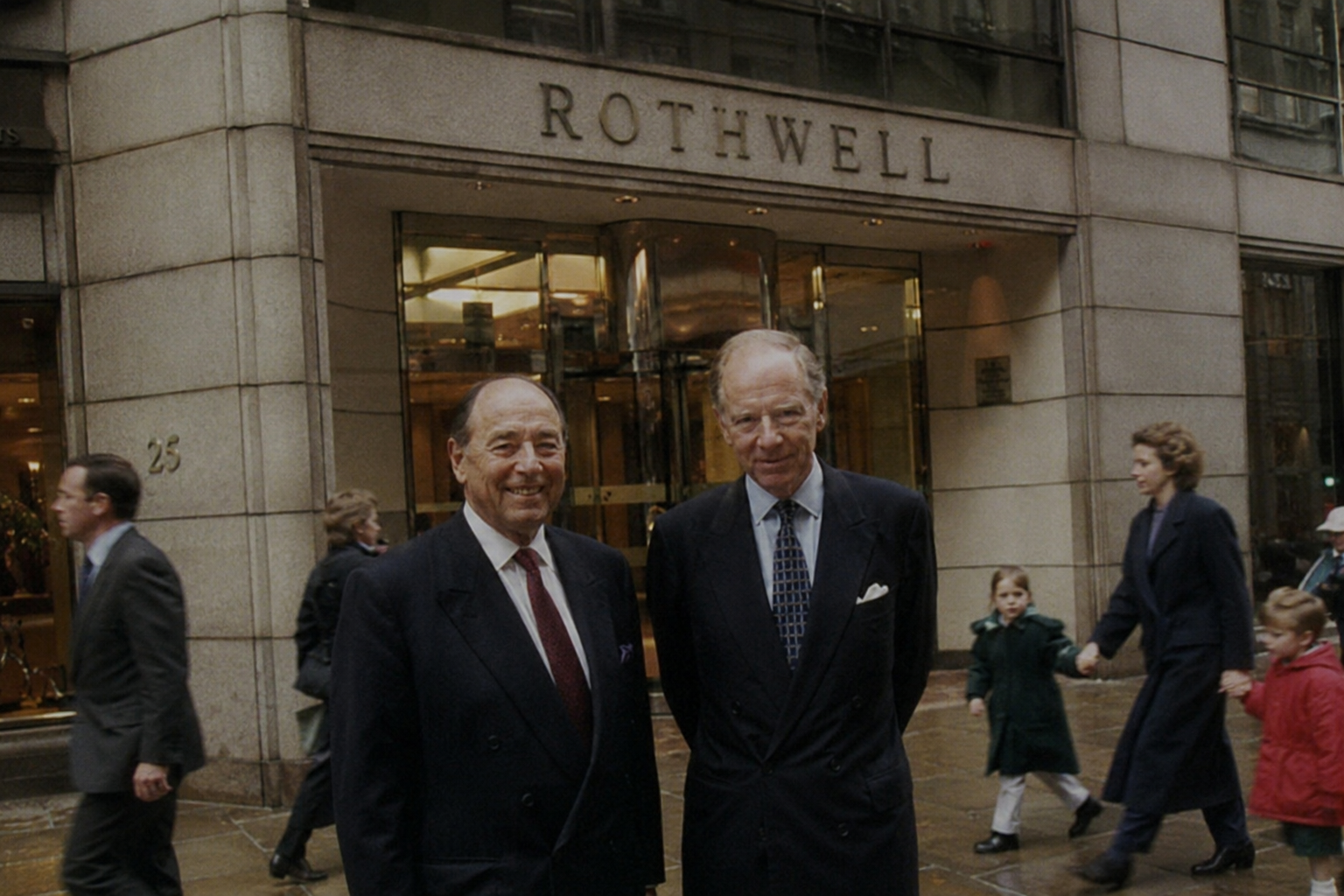 Two men in suits standing outside the Rothwell building on a rainy day, with other pedestrians walking by.