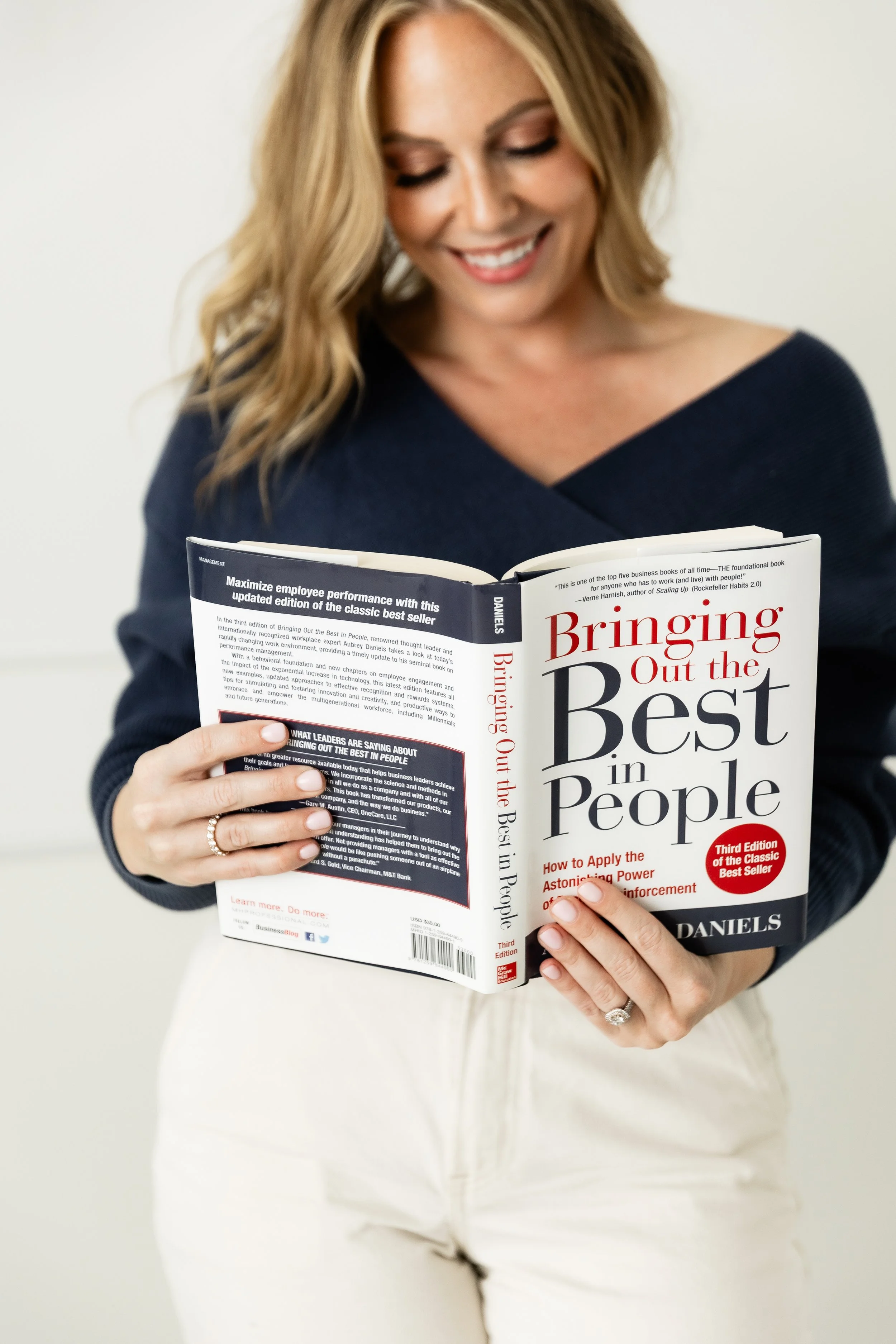 A woman with shoulder-length blonde hair, wearing a navy blue top, is smiling and holding a book titled 'Bringing Out the Best in People' in front of a plain light-colored background.