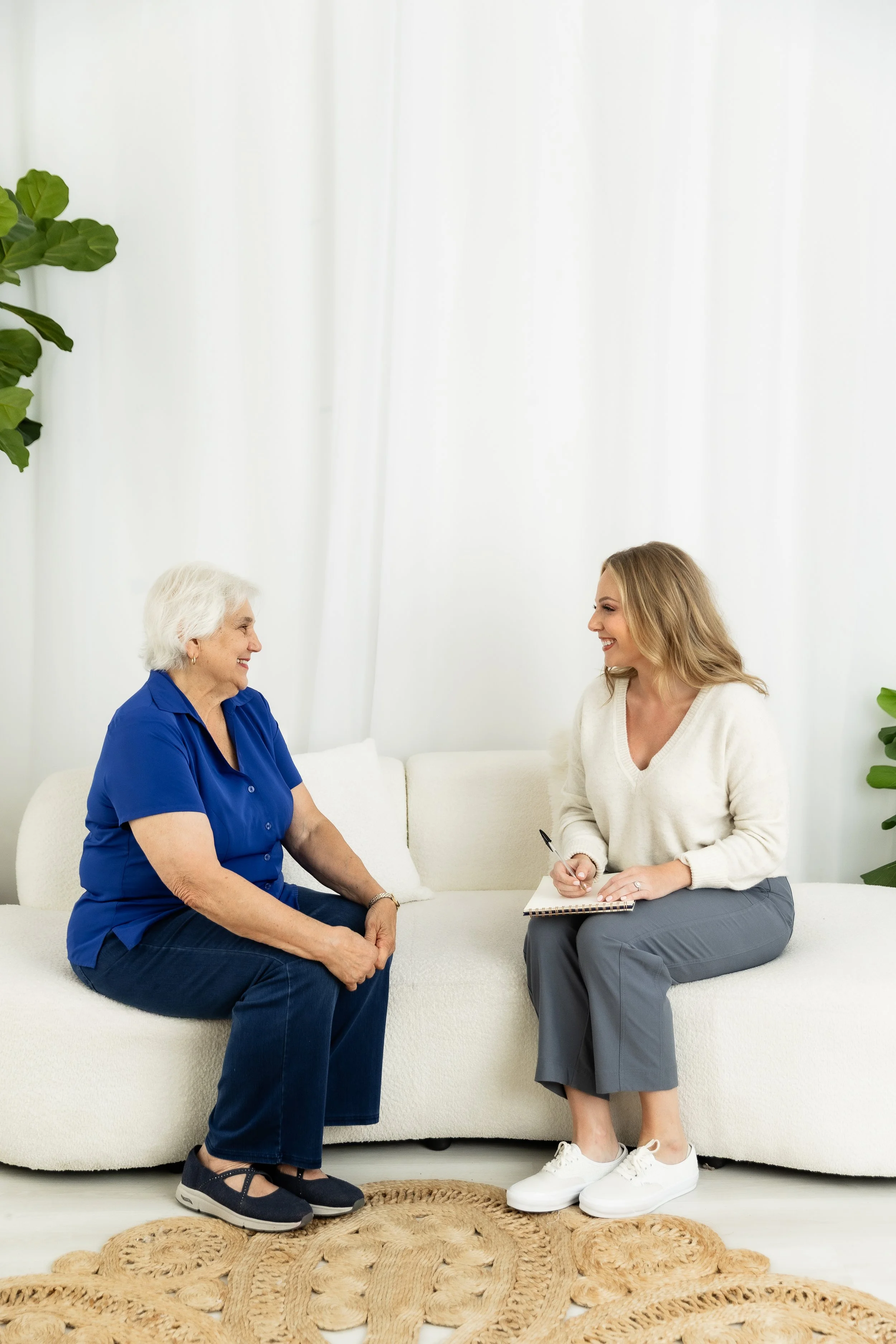 Two women, one older with gray hair in a blue shirt and the other younger with blonde hair in a white sweater, are sitting and conversing in a bright, minimalist room. The younger woman is holding a pen and notebook, smiling, while the older woman is attentively listening.