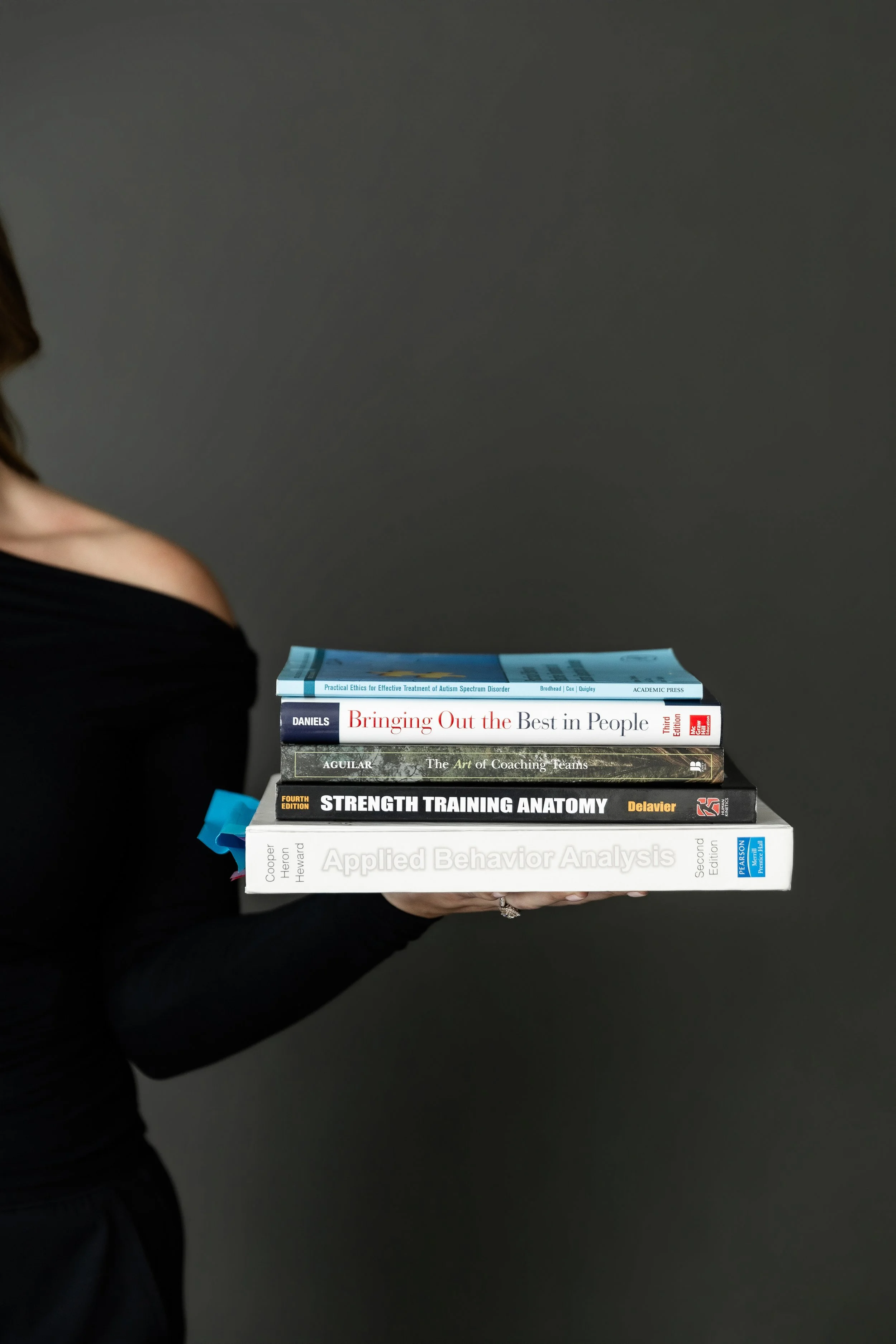 A person wearing black clothing is holding a stack of six books against a plain dark background. The books are stacked horizontally with their spines facing outward, displaying titles related to psychology, coaching, strength training, and behavior analysis.