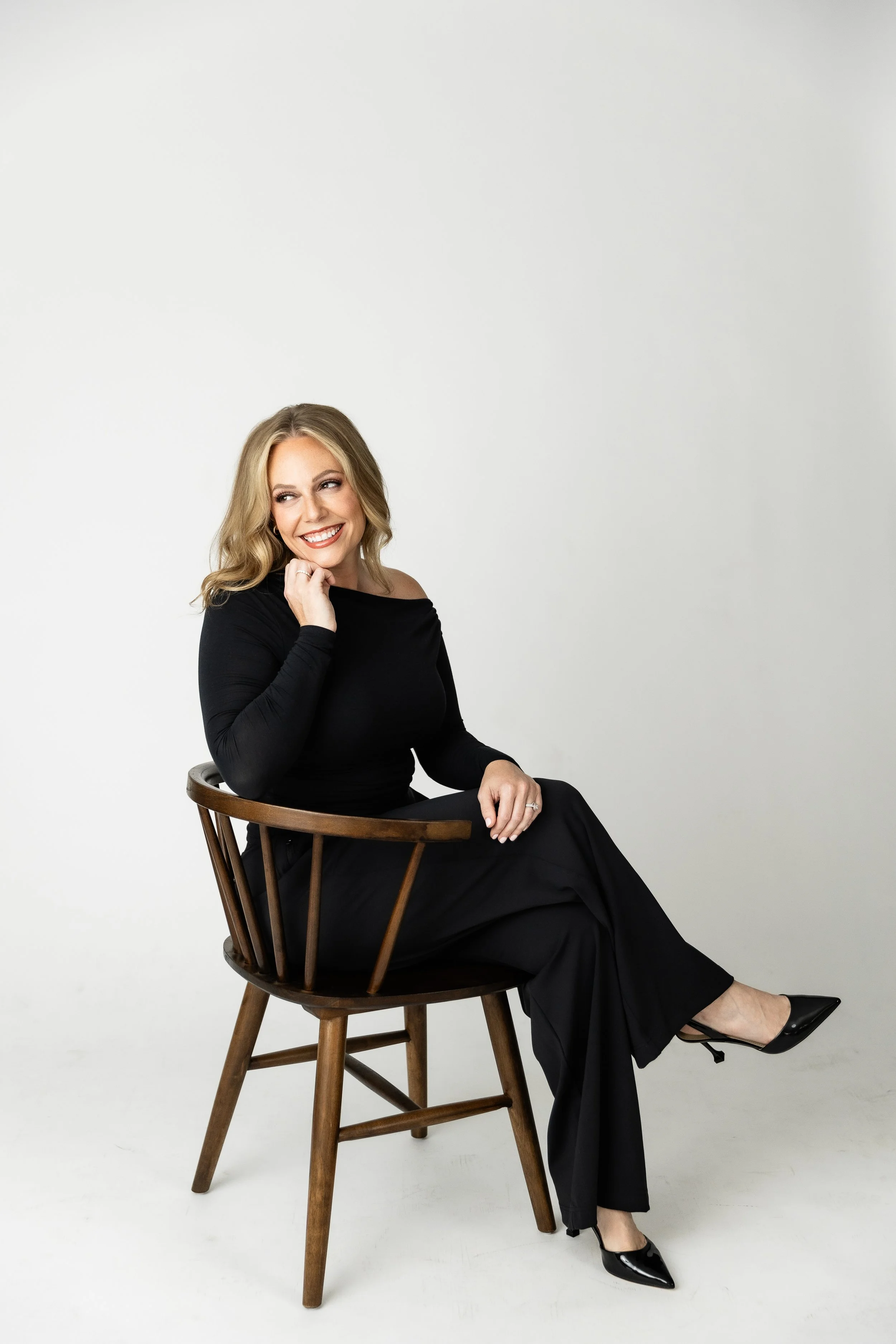 A woman with blonde hair wearing a black off-shoulder top and black pants, sitting sideways on a wooden chair, smiling and looking to her left, with one hand resting near her chin, set against a plain light background.