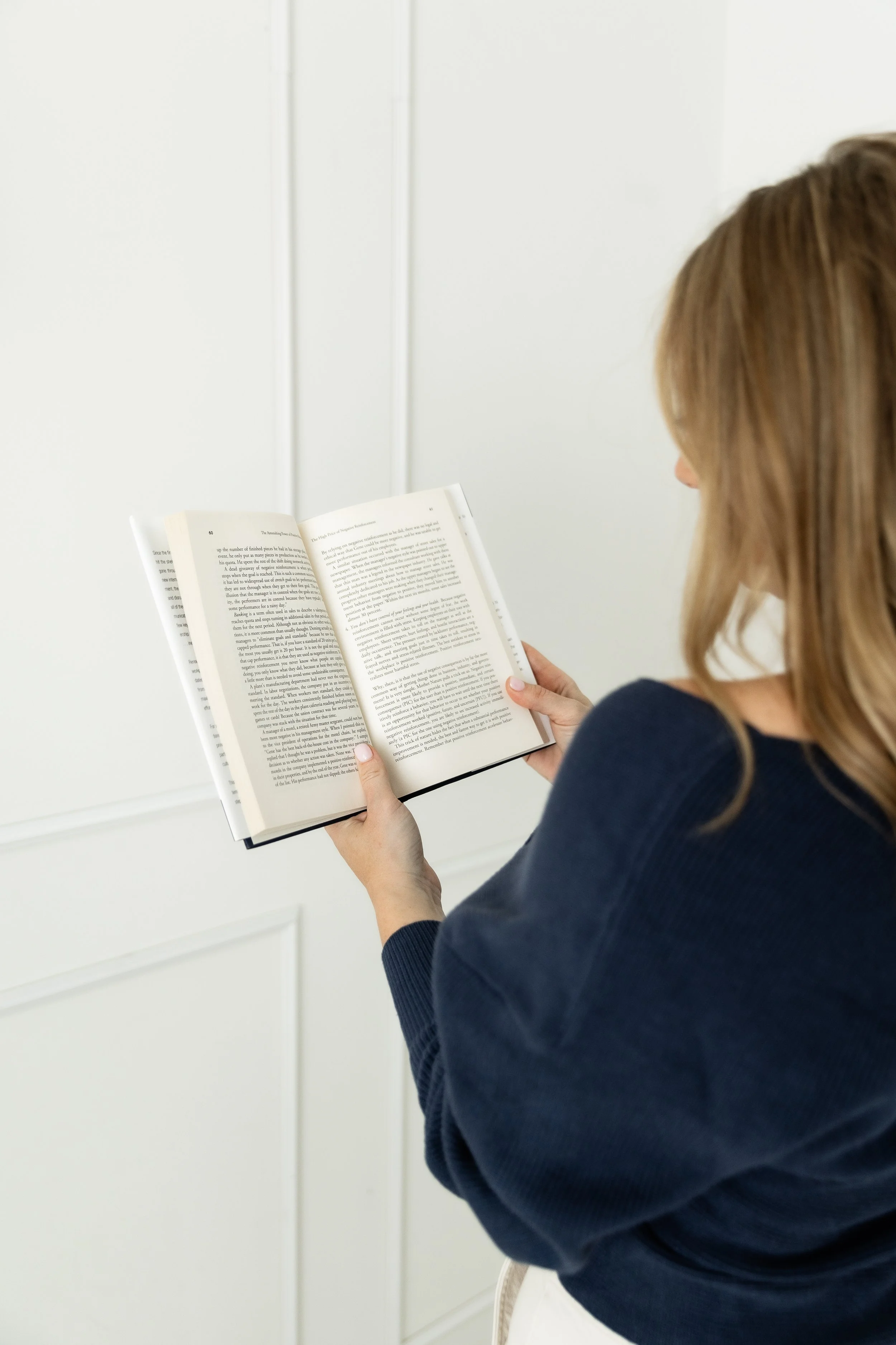 A woman with brown hair reading a hardcover book in a room with white walls.