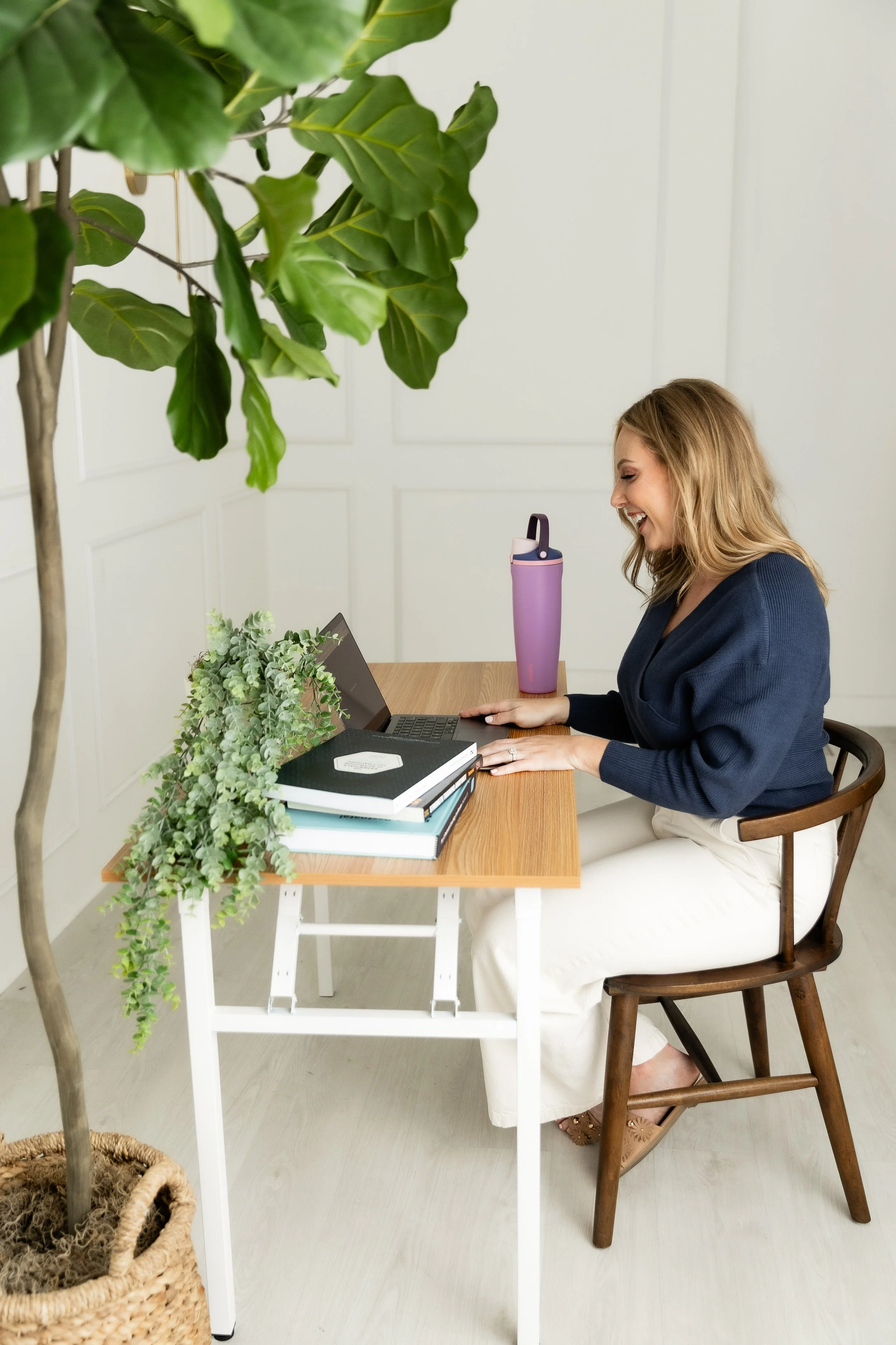 A woman with blonde hair sitting at a wooden desk, smiling while looking at a laptop, with a purple water bottle, books, and plants on the desk, and a green plant in the foreground.