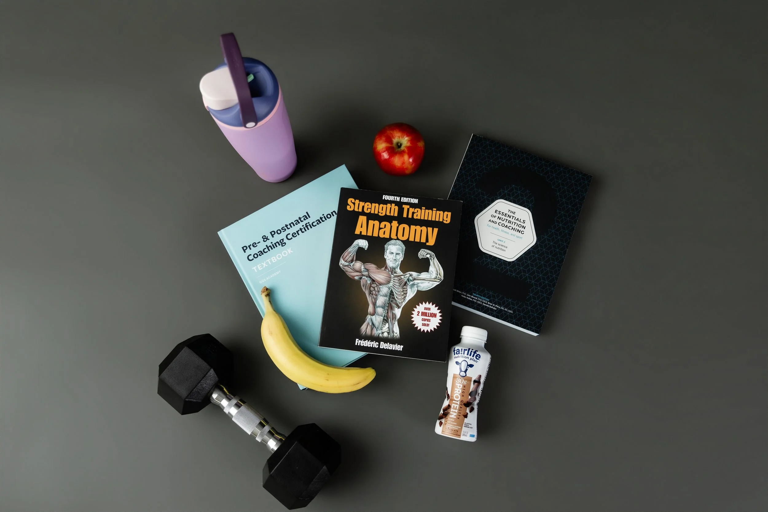 A flat lay of fitness and nutrition items on a gray background, including a black dumbbell, a purple water bottle, a banana, two books about strength training and nutrition, a red apple, a bottle of protein supplement, and a white bottle of dietary supplement.