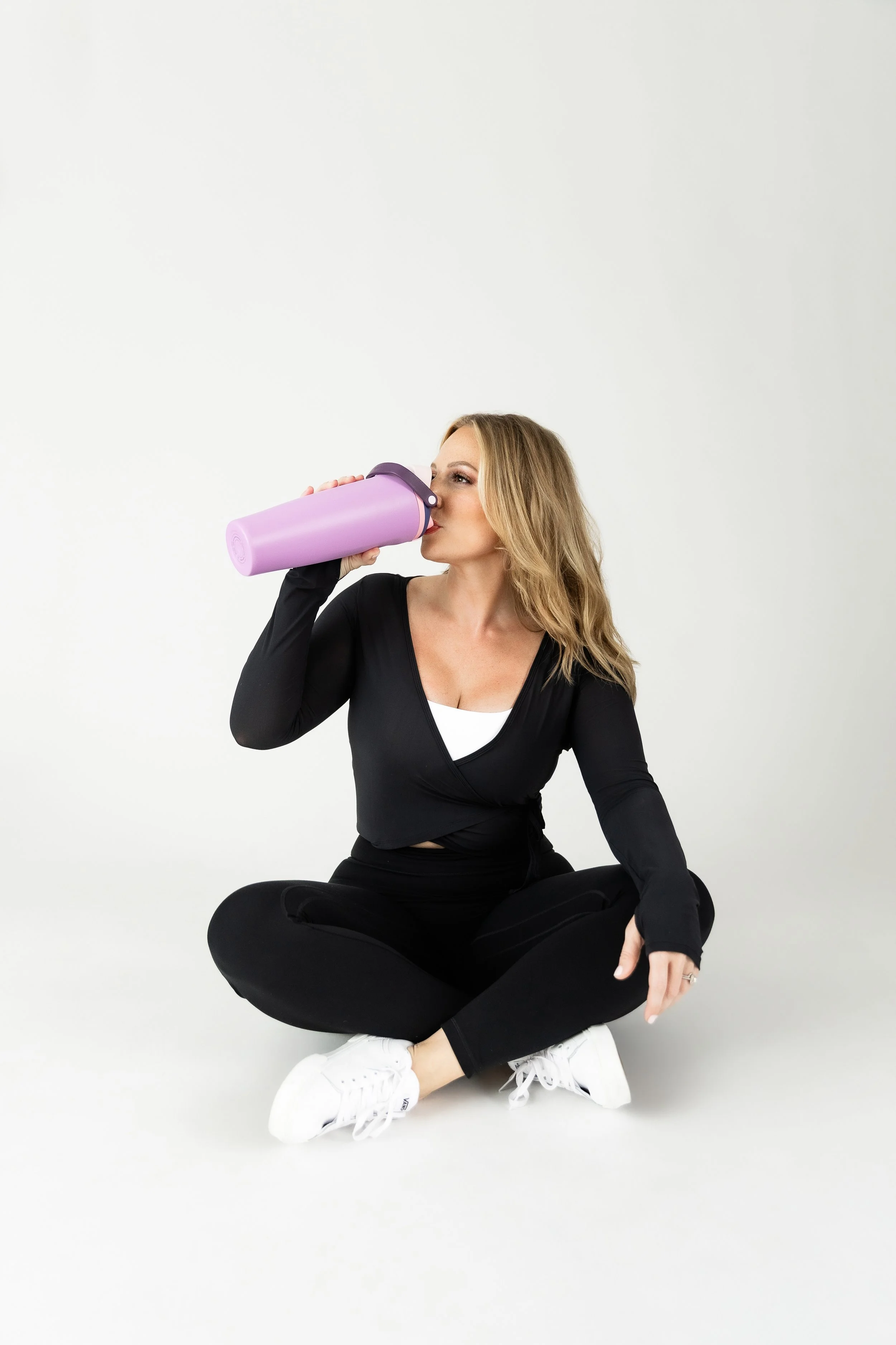 A woman with long blonde hair, wearing a black athletic top and black leggings, sits cross-legged on the floor, drinking from a purple water bottle.