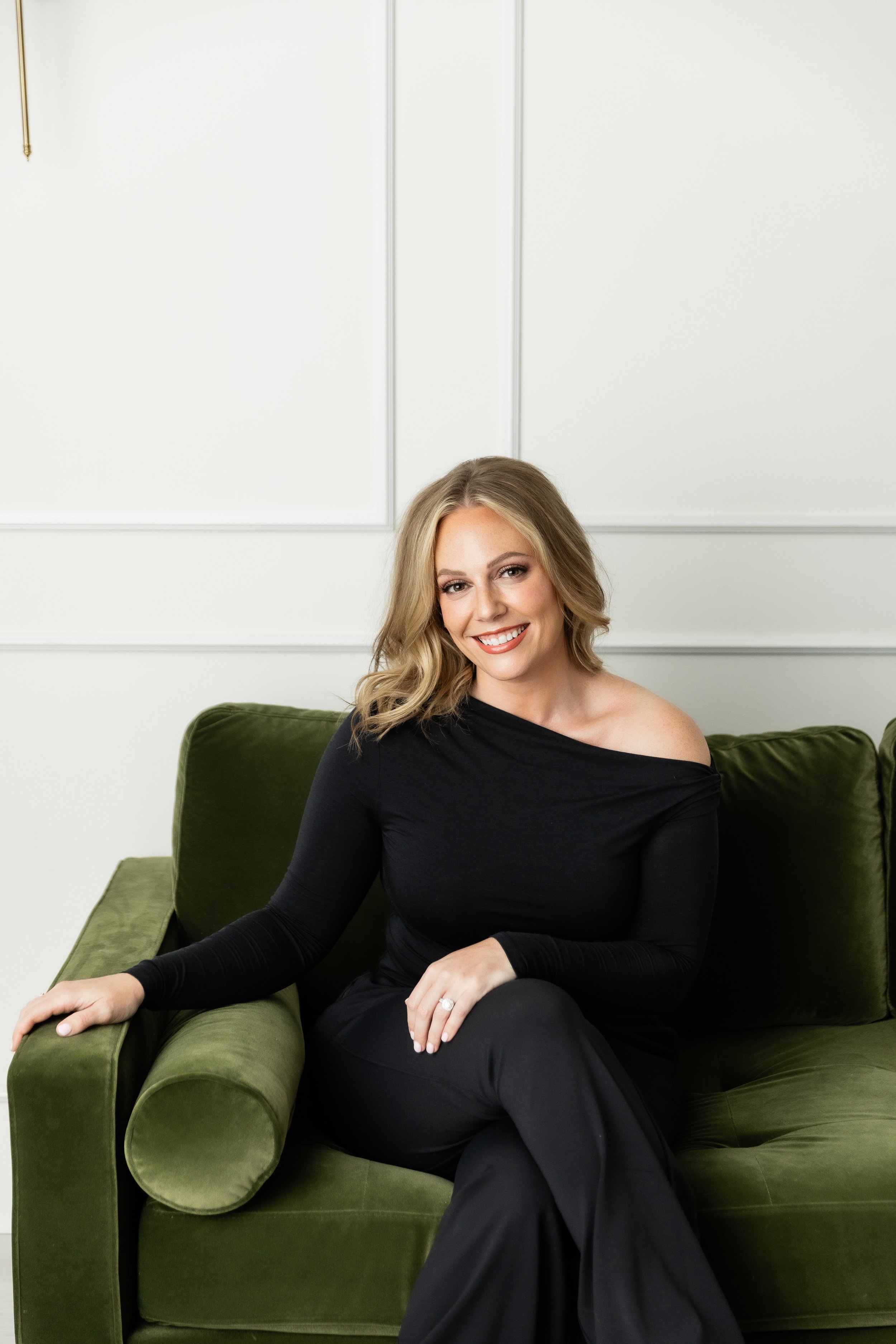 A woman with blonde hair and a black top sitting on a green velvet couch indoors.