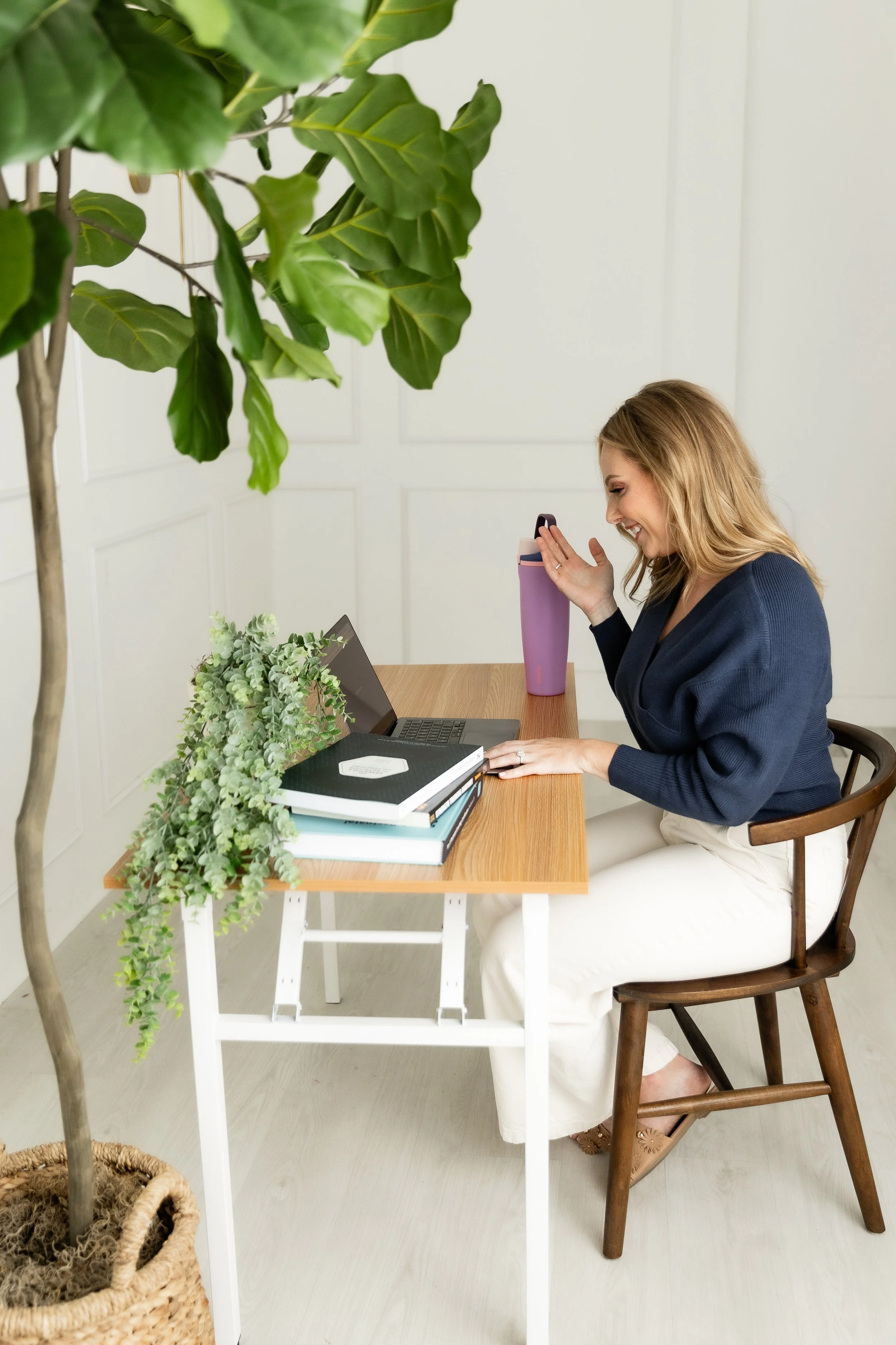Woman sitting at a desk with a laptop, books, a purple travel mug, and plants, smiling and looking at her phone in a bright room.