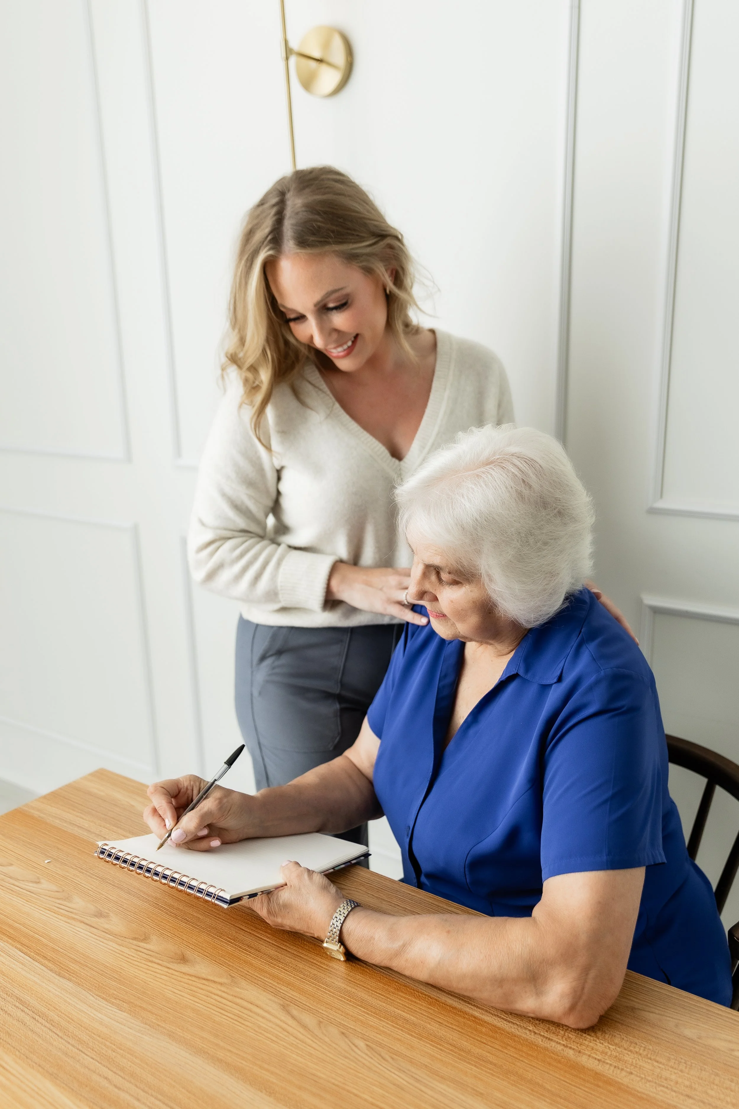 A woman in a beige sweater standing next to an elderly woman in a blue shirt who is signing a document on a wooden table.