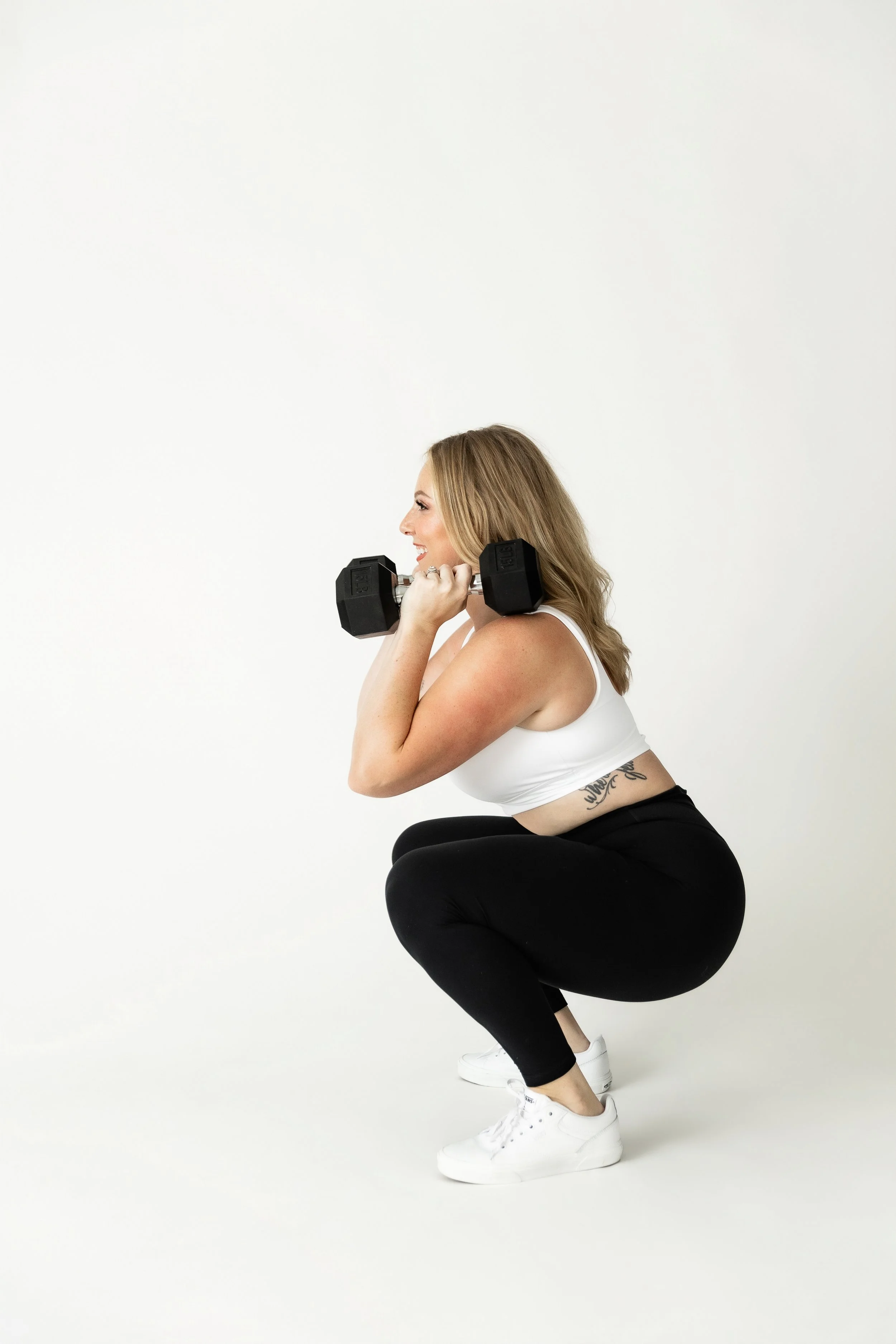 A woman squatting while holding a dumbbell in each hand, wearing a white tank top, black leggings, and white sneakers, with a tattoo on her side, against a plain white background.