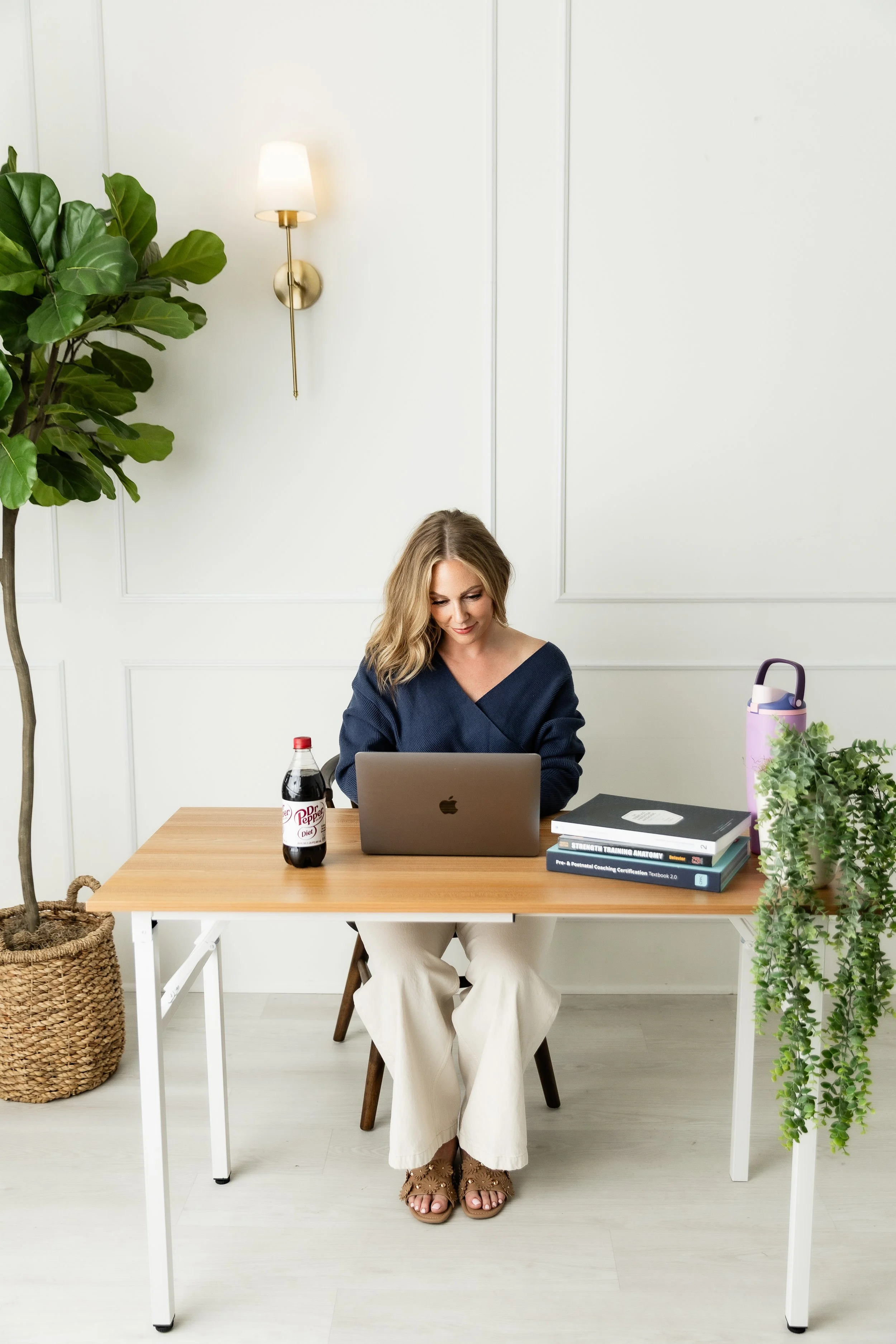 A woman sitting at a wooden table using a silver MacBook laptop in a bright room. There are books, a bottle of Diet Dr Pepper, and plants on the table, with a large potted plant to the left, and a woven basket with a plant on it. A wall sconce and white paneled wall are in the background.