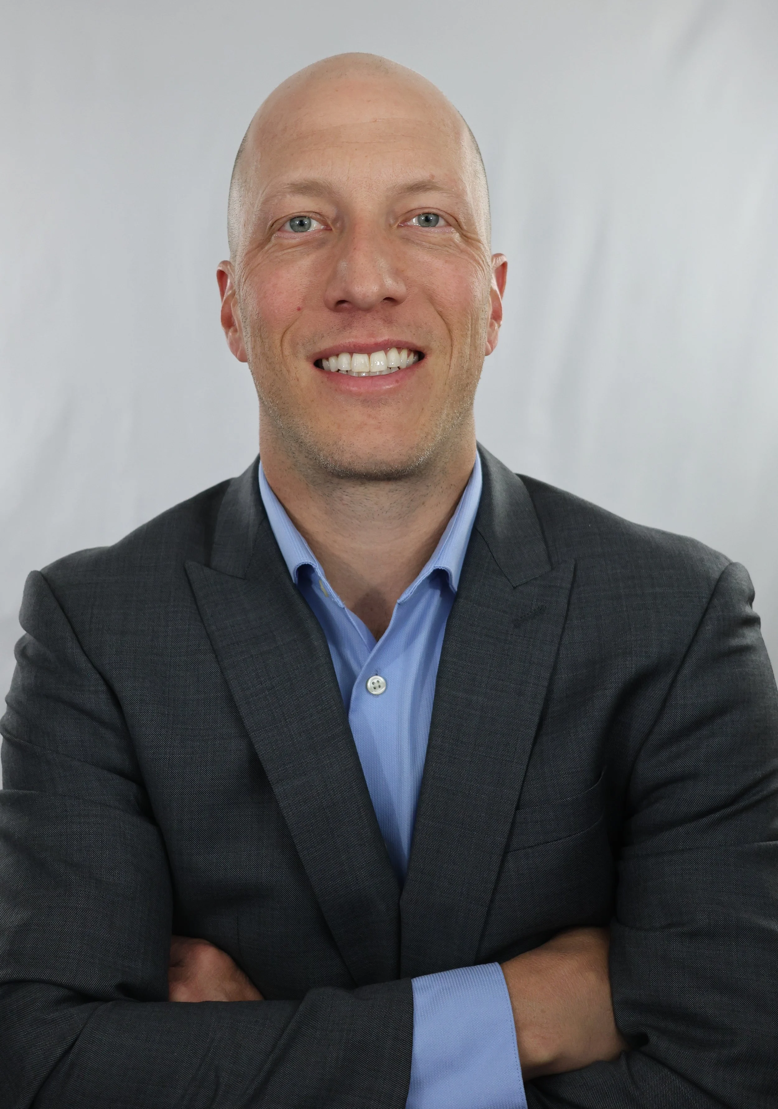 Portrait of a man with a bald head and blue eyes, wearing a dark suit and light blue dress shirt, smiling with arms crossed against a plain light gray background.