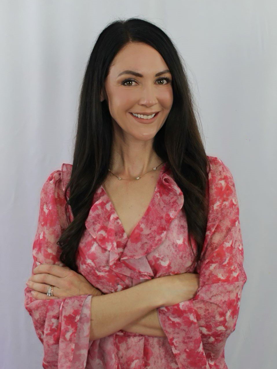 Smiling woman with long dark hair wearing a pink patterned dress standing against a light background.
