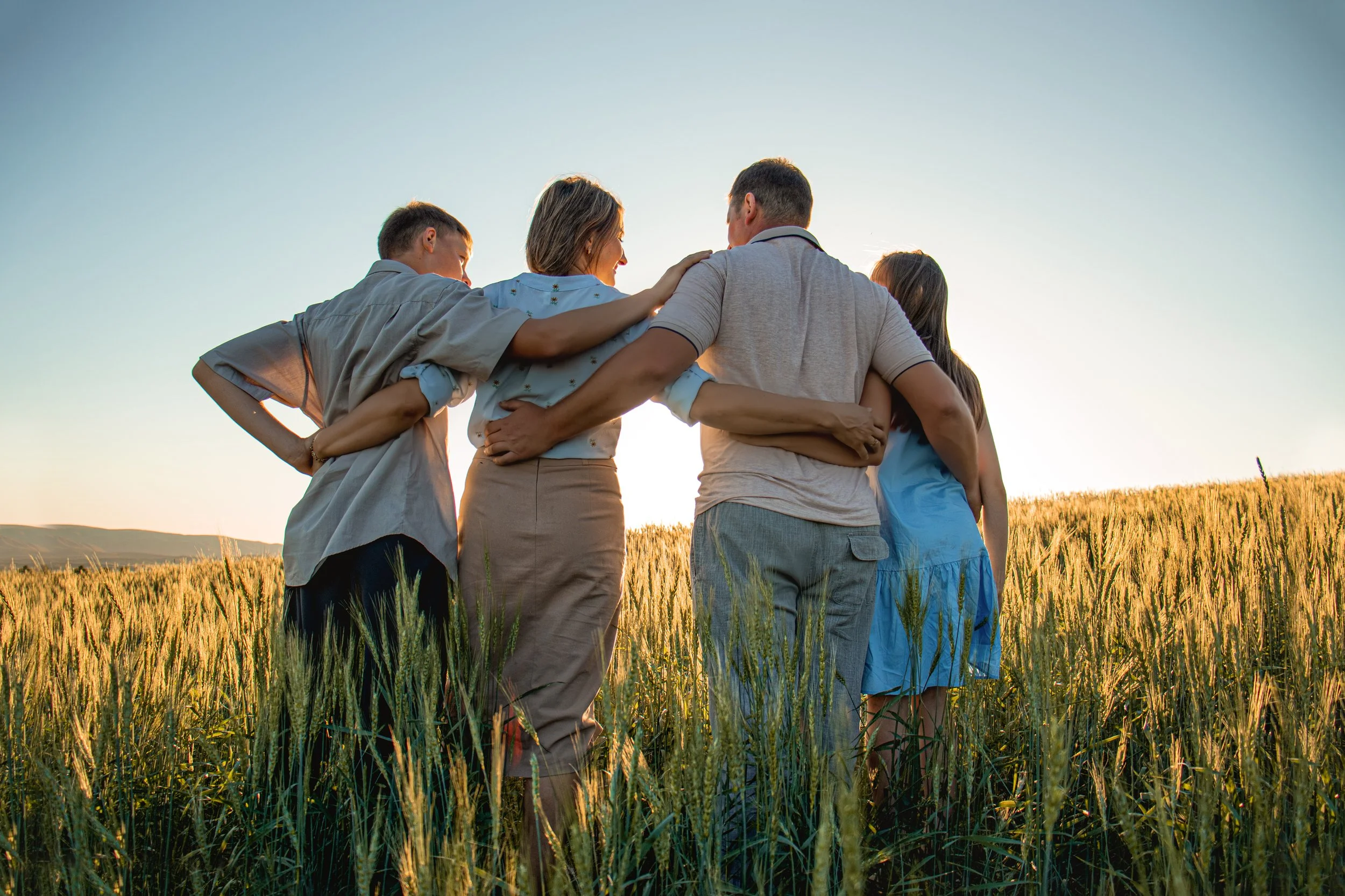 Group of five people standing together in a field of tall wheat during sunset, with their arms around each other, facing each other and smiling.