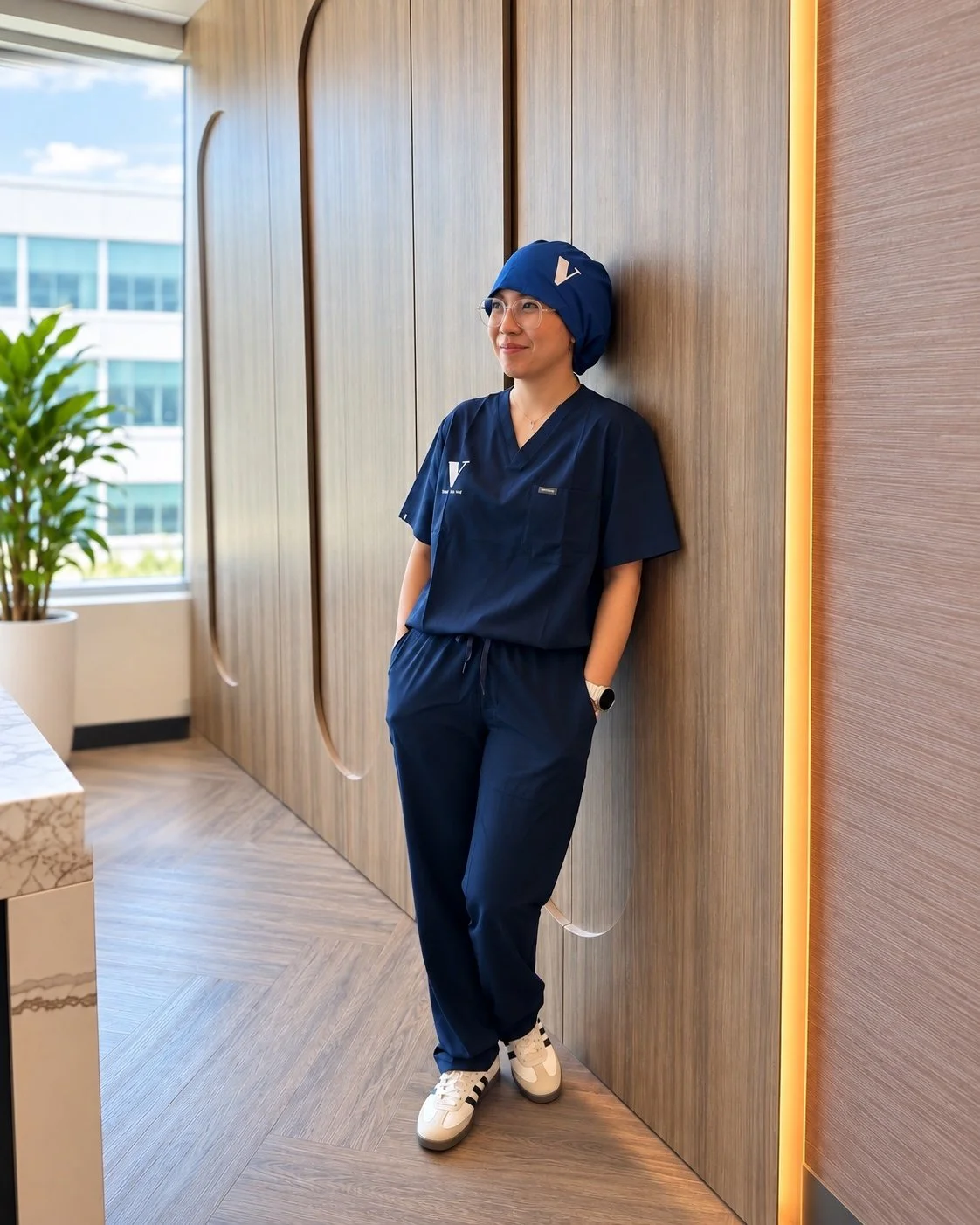 A person in a blue medical uniform, hat, and glasses, leaning against a wooden wall with hands in pockets in a modern office setting.