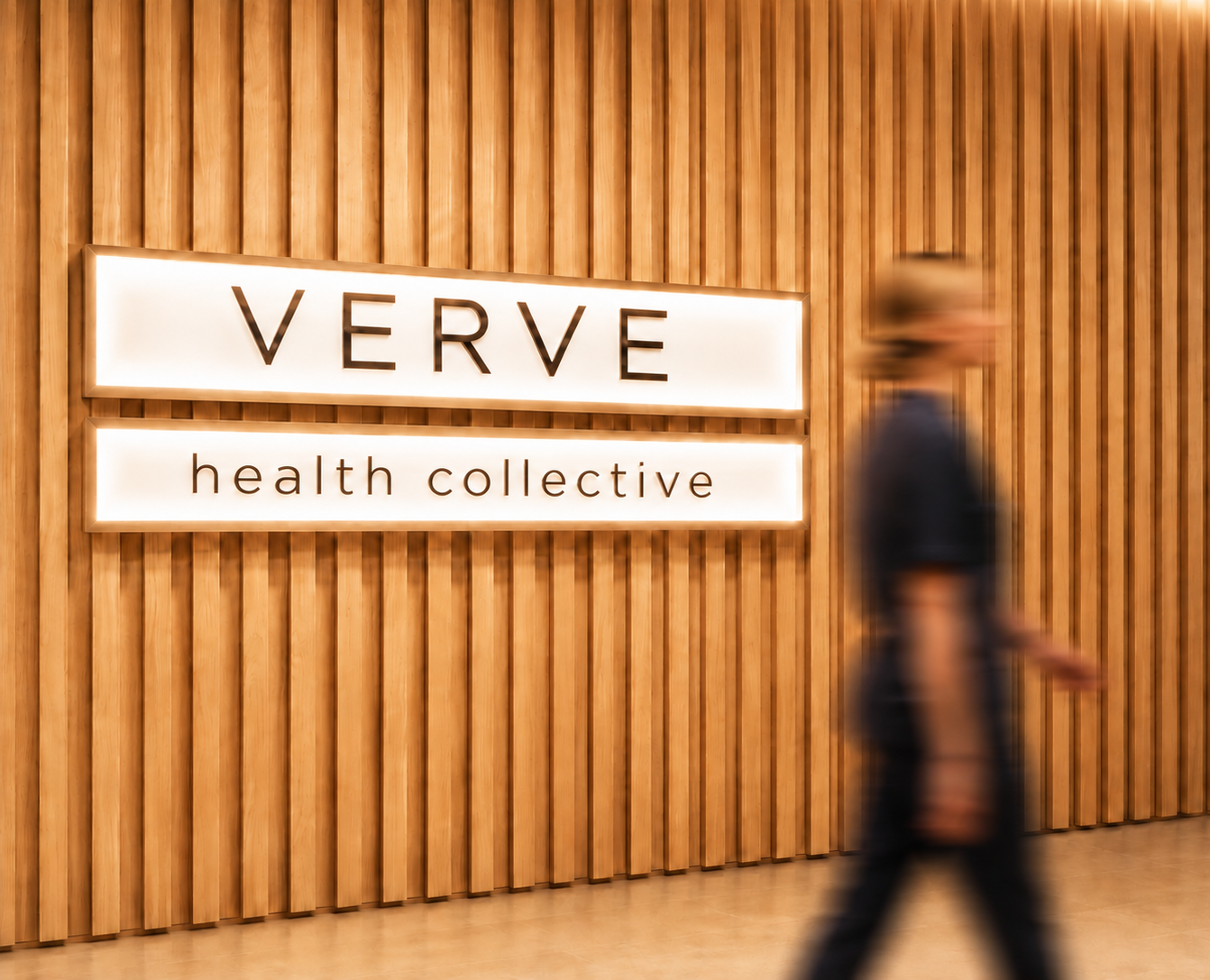 Blurry person walking past a wooden wall with illuminated sign that reads 'VERVE health collective'.