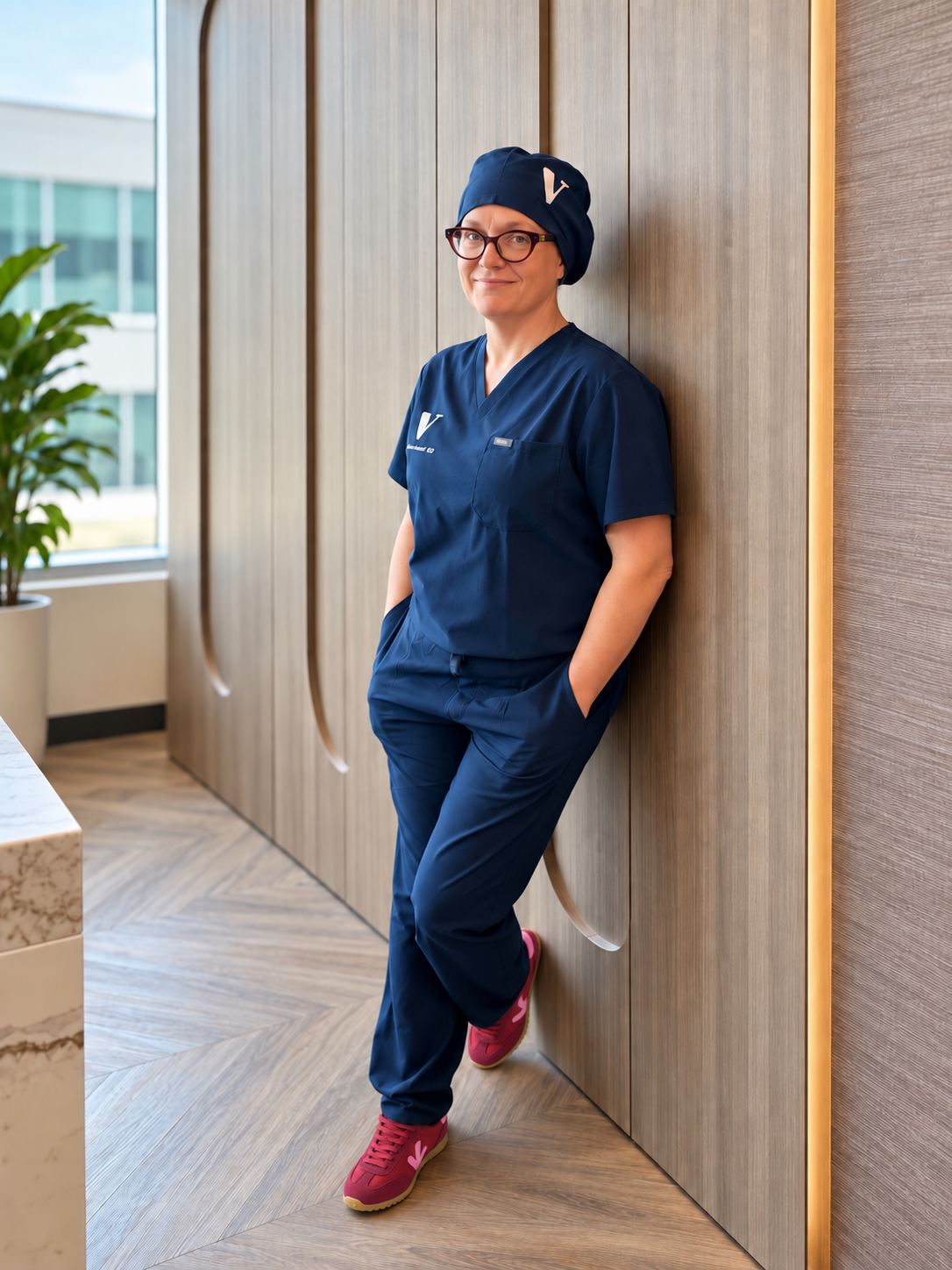 Dr Liane Belland - A woman wearing navy blue medical scrubs, red glasses, and pink sneakers, leaning against a wooden wall in a modern indoor setting.