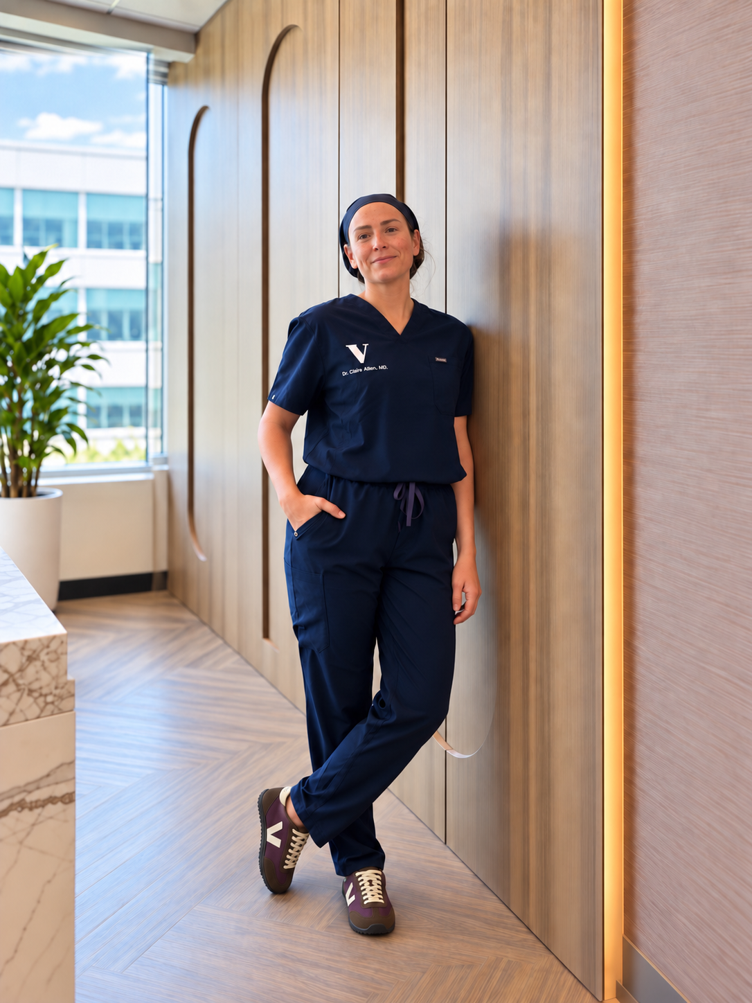 Dr Claire Allen - A woman in navy medical scrubs, standing in a modern office hallway with wood-paneled walls and a large window, smiling with hands in pockets.